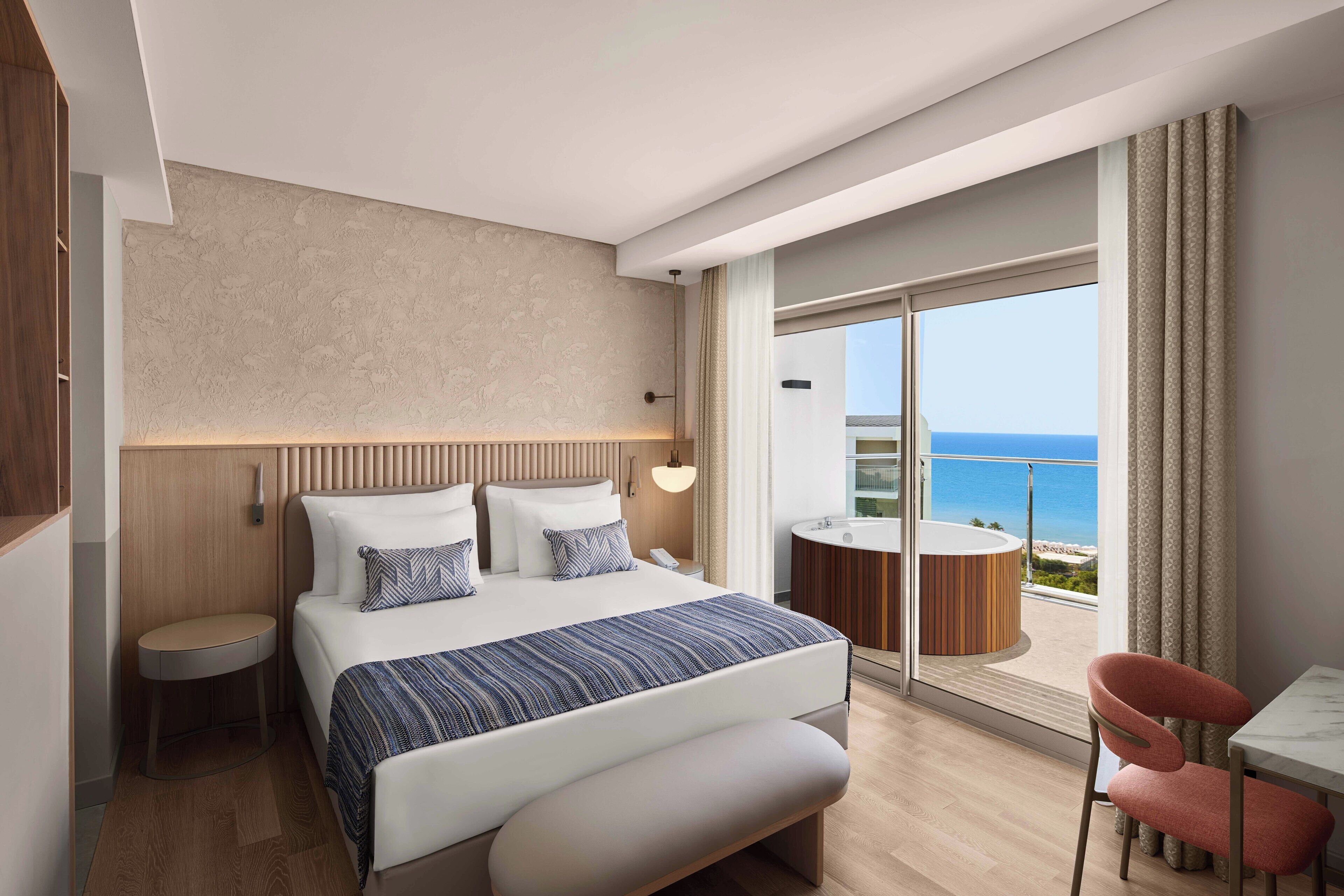 Une chambre d’hôtel moderne avec un grand lit, une petite table ronde, une chaise et une vue sur l’océan, visible à travers une porte-fenêtre coulissante.