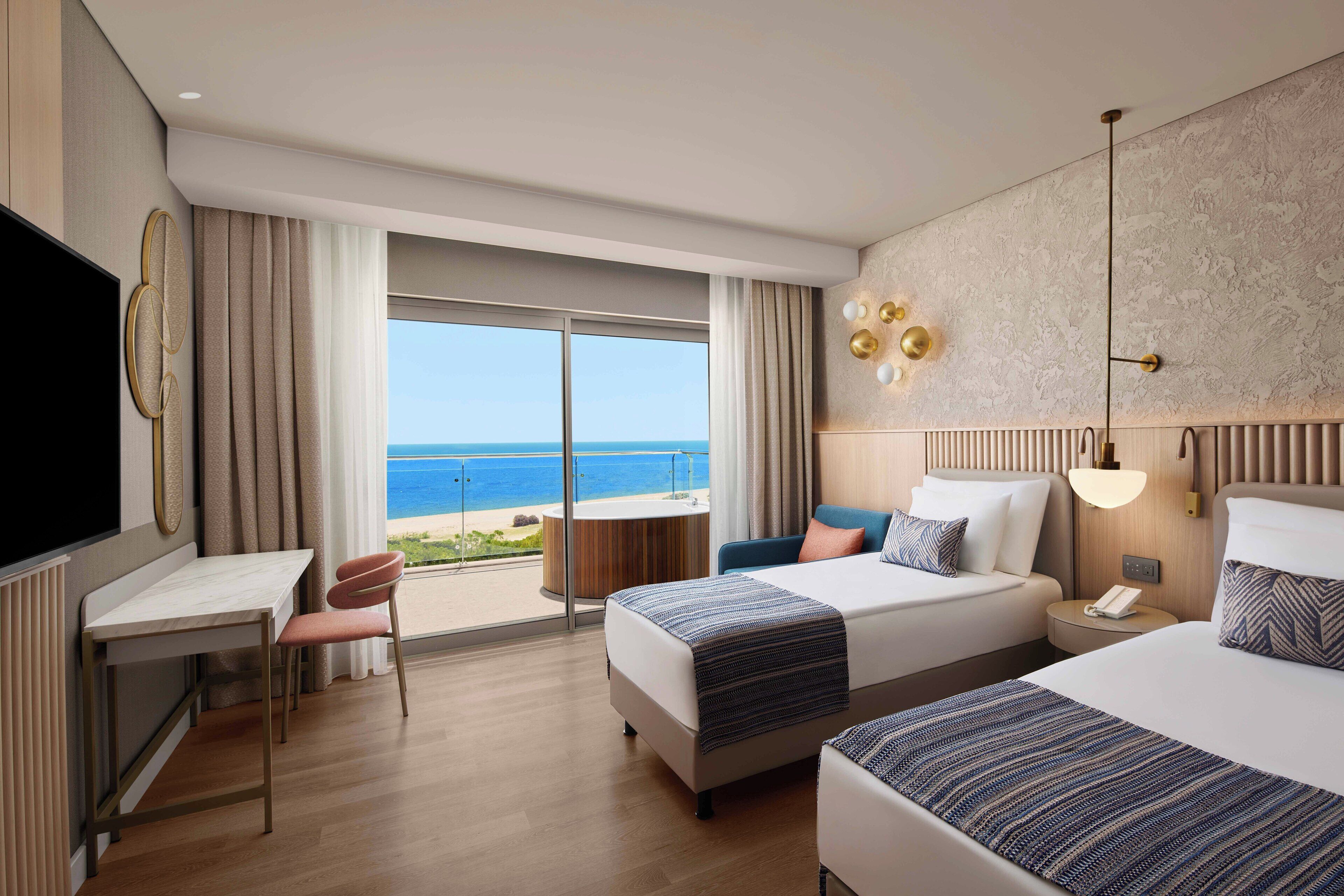 A hotel room with two beds, a desk, a chair, and a TV. The room has a view of the ocean.
