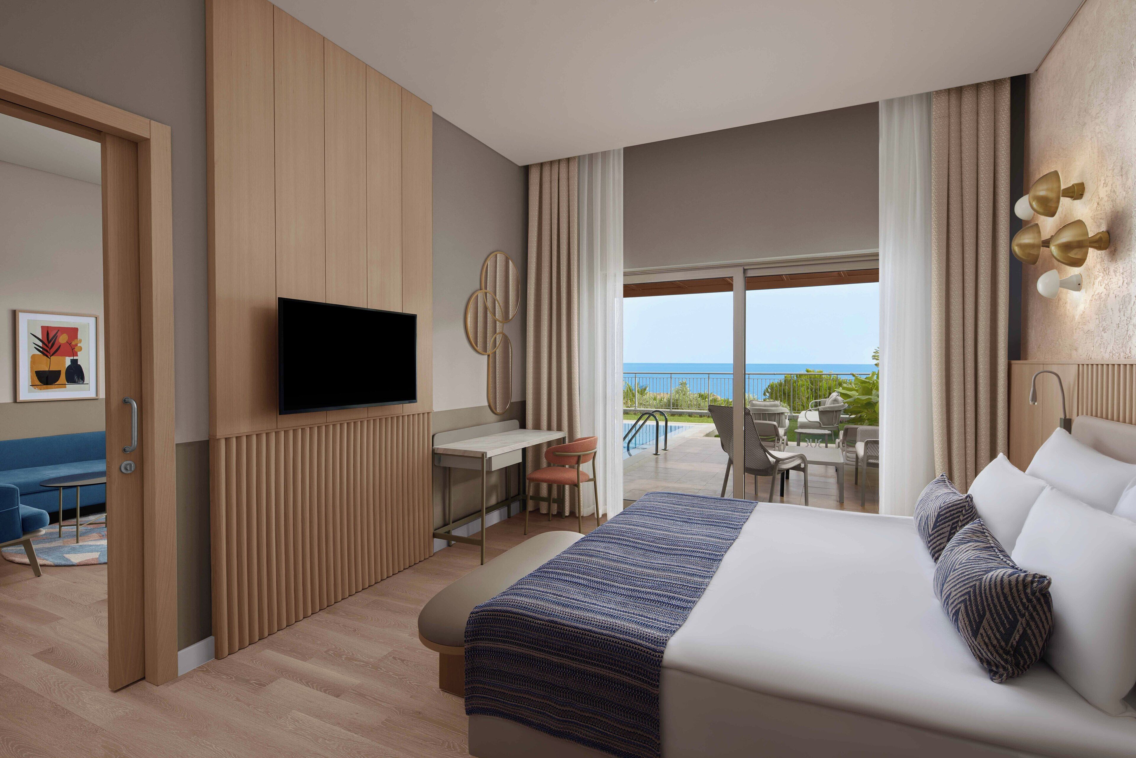 A modern hotel room with a bed, a desk, and a TV. The room has a view of the ocean.