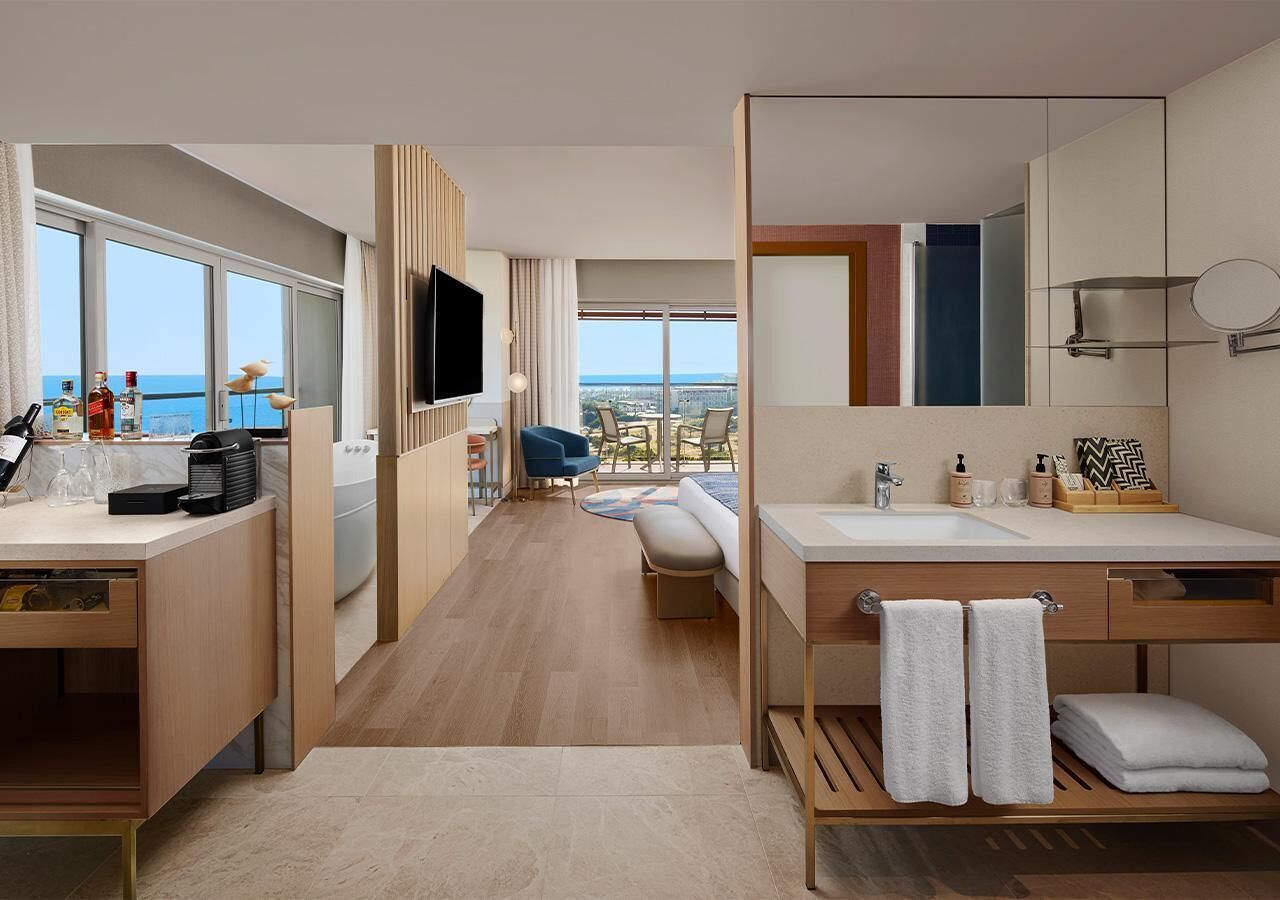 A modern hotel room with a large window, a flat-screen TV, a desk with a coffee machine, a sofa, and a bathroom with a sink and mirror.