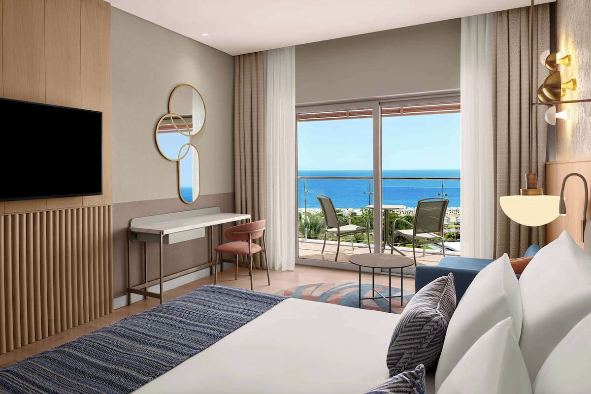 A modern hotel room with a large bed, a desk, a flat-screen TV, and a balcony with a view of the sea.