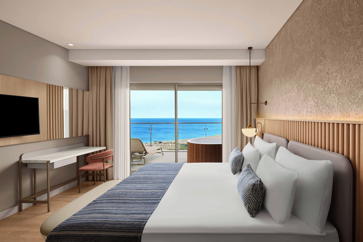 A modern hotel room with a large bed, a desk, a chair, a TV, and a balcony with a view of the sea.
