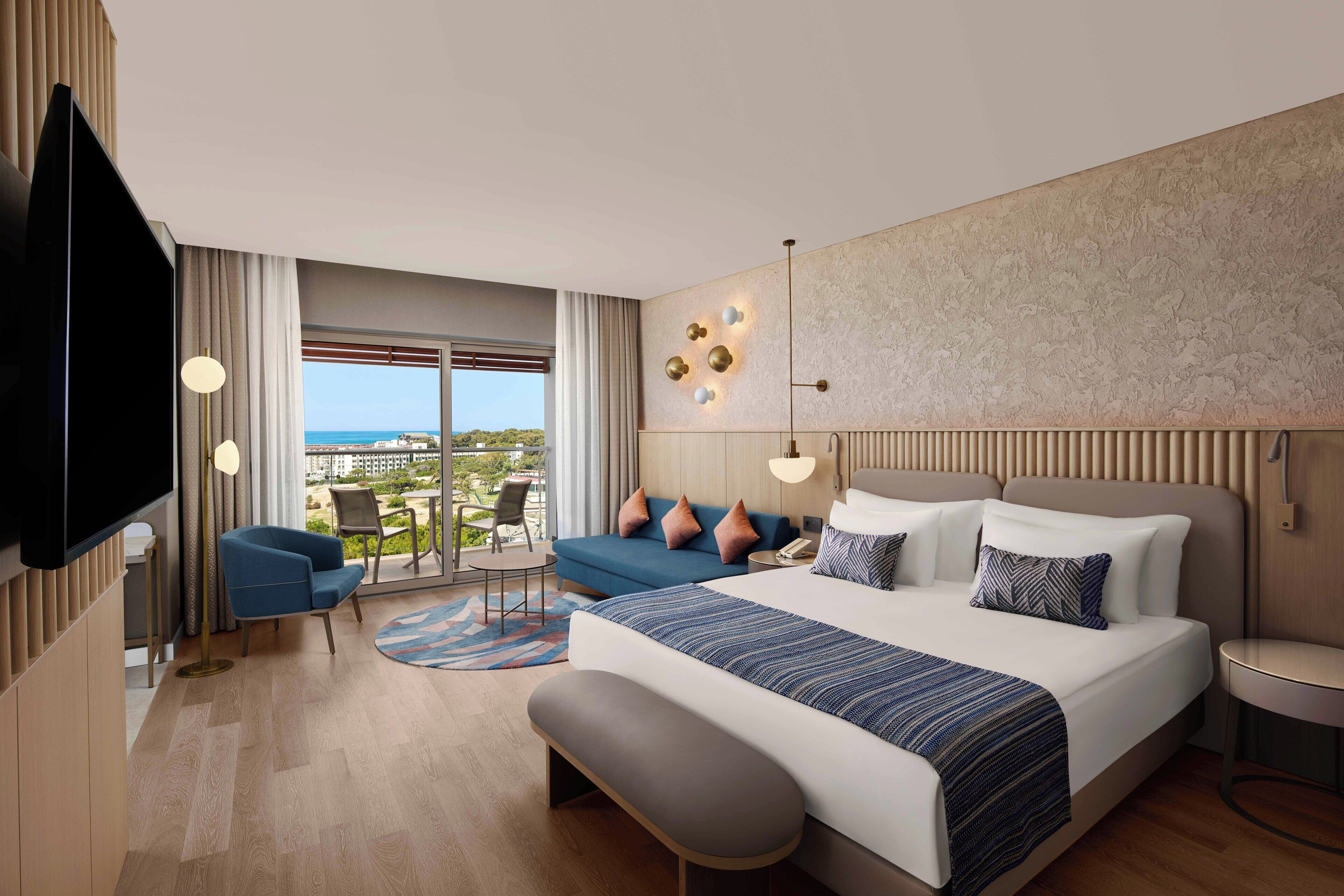 A modern hotel room with a large bed, a sofa, a TV, and a balcony with a view.
