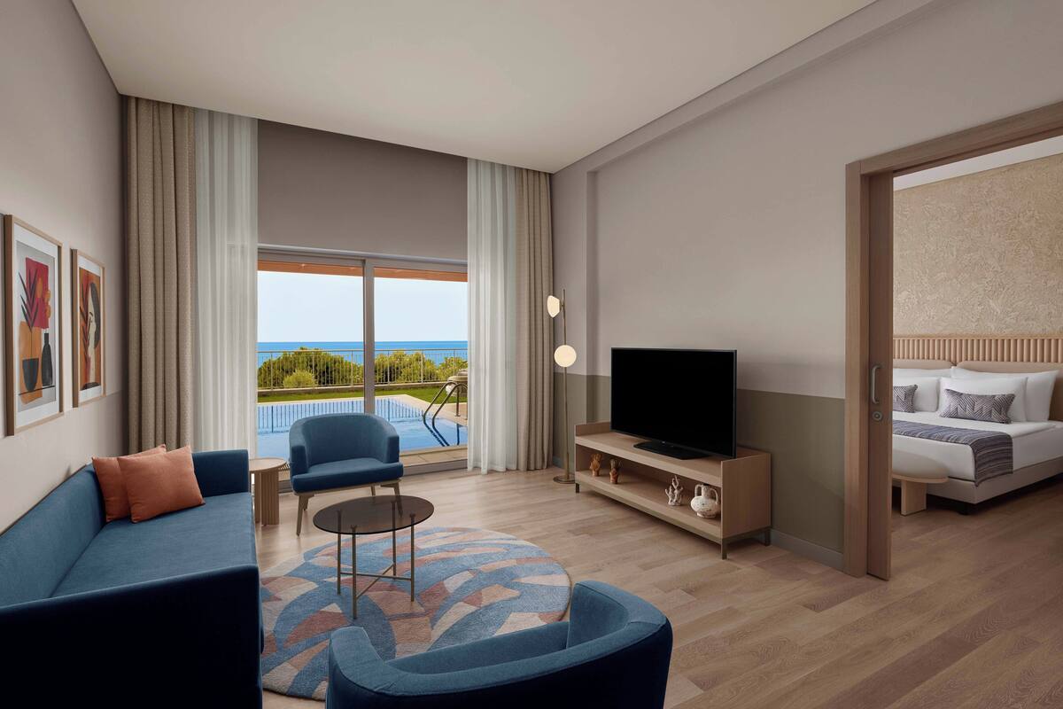 A modern hotel room with a sofa, a television, a coffee table, and a view of the pool and greenery.