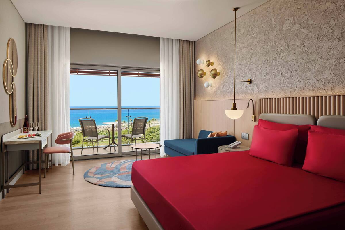 A hotel room with a large bed, a desk with a chair, a small table with a chair, and a view of the ocean through large windows.