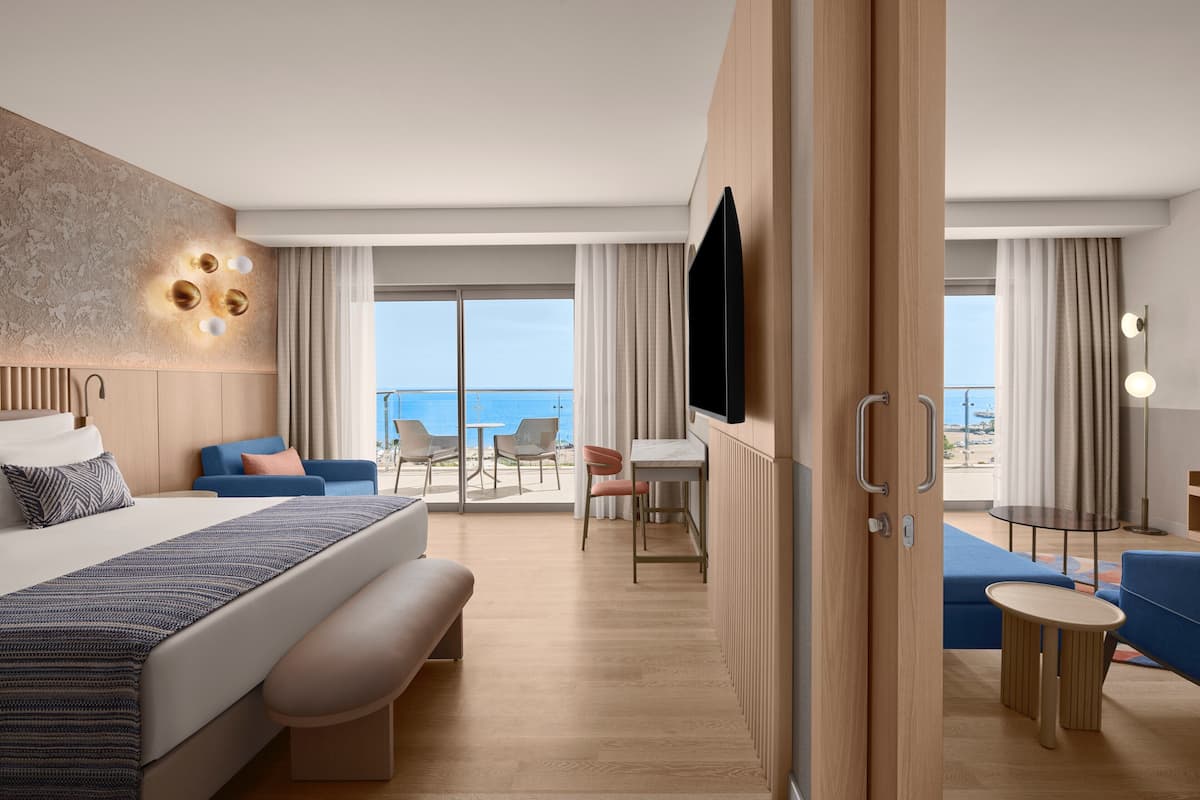 A modern hotel room with a large bed, a desk, a chair, a small table, and a TV. The room has a view of the ocean.