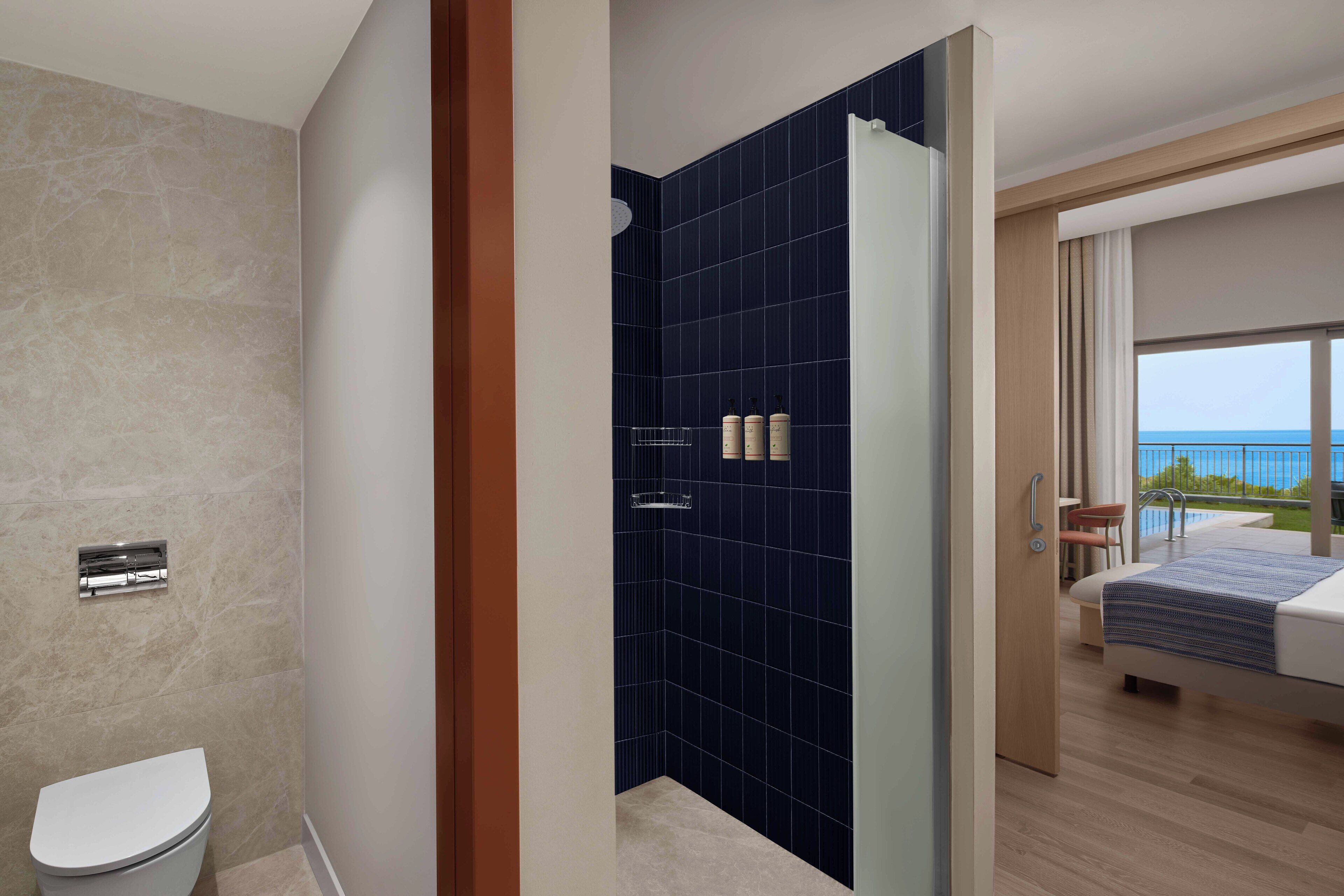 Une salle de bain moderne avec des toilettes blanches, une douche aux carreaux bleu foncé et une vue sur un balcon équipé d’un lit et d’une chaise.