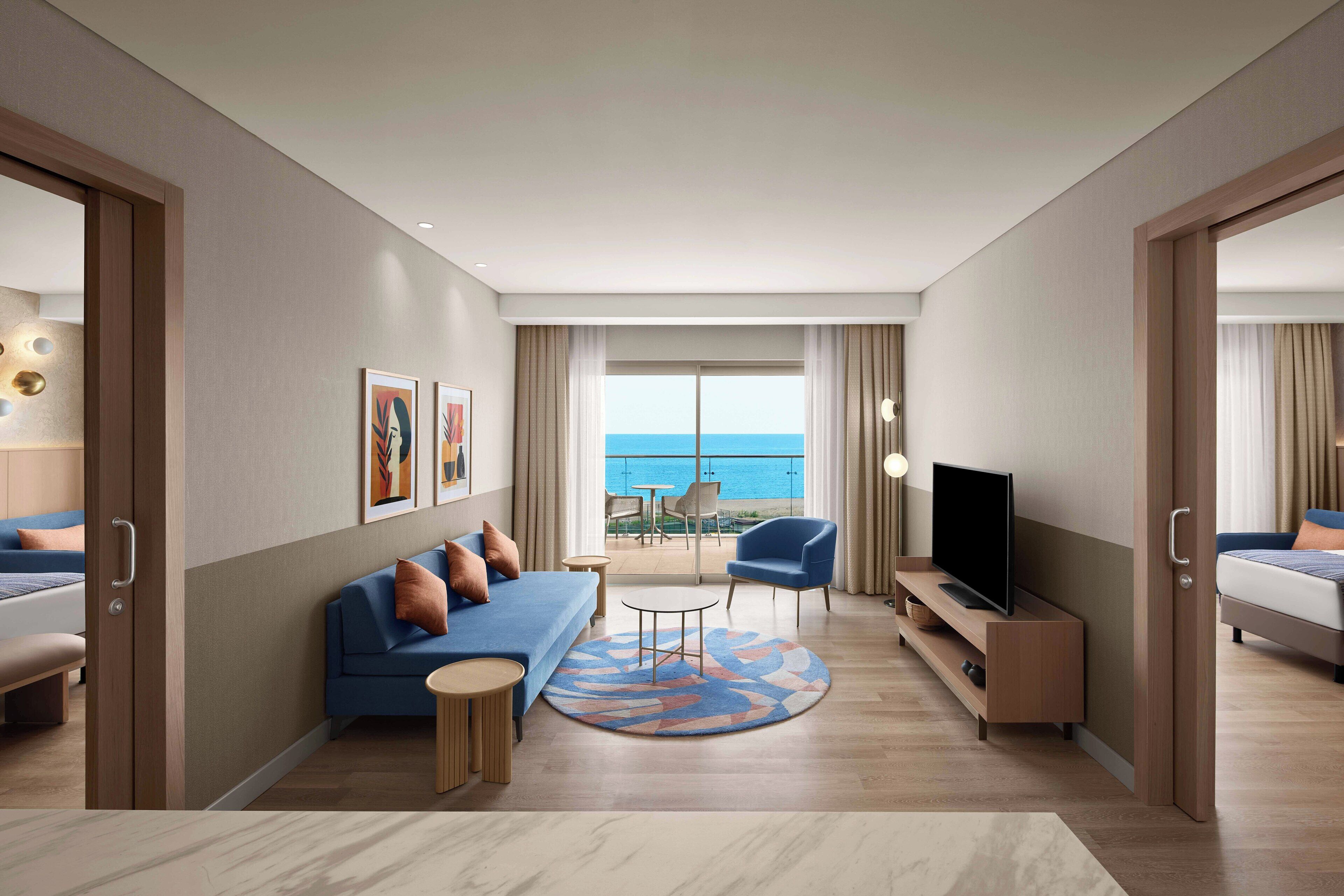 A modern hotel room with a large balcony, a sofa, a TV, and a dining area.