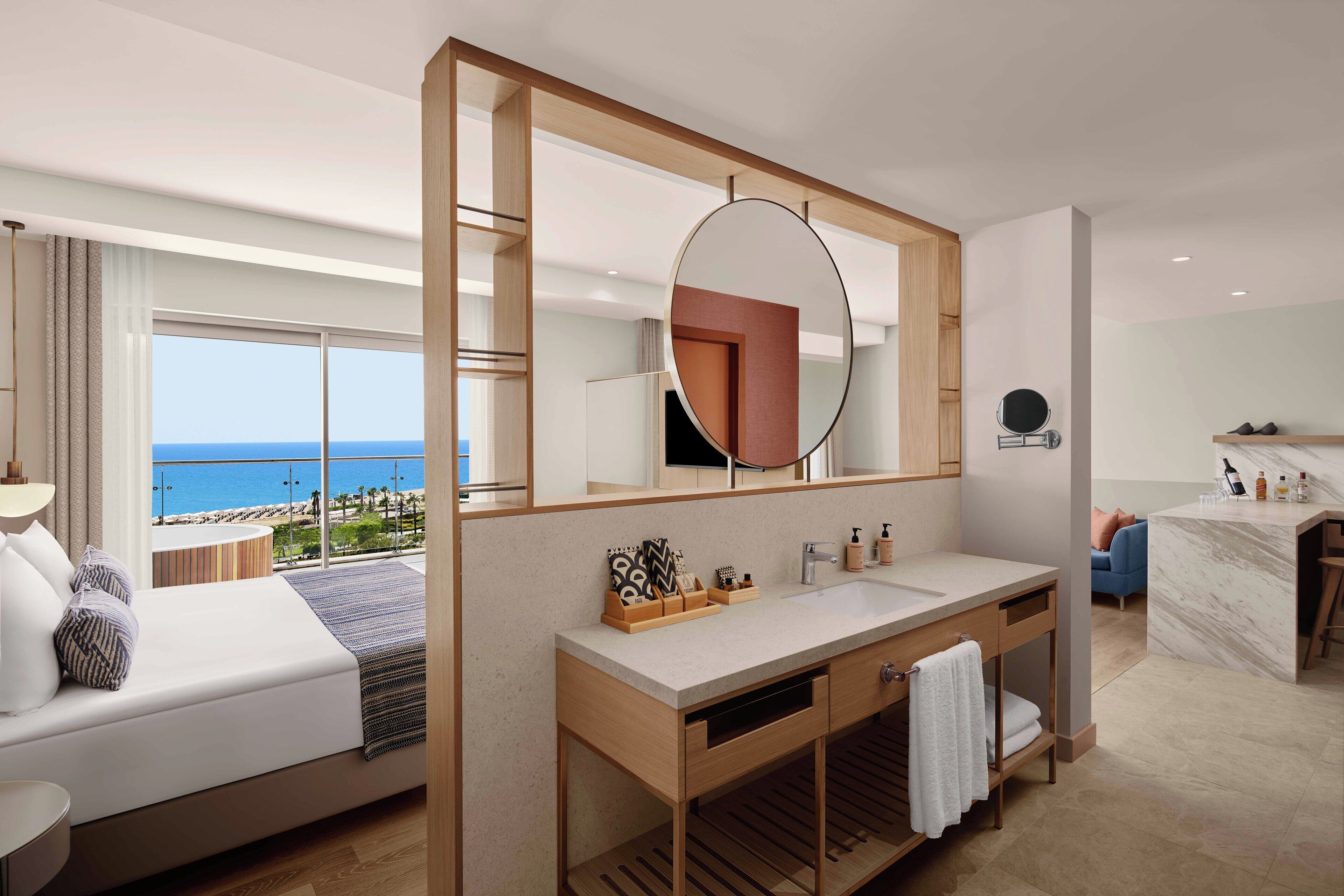 A modern hotel room with a large bed, a wooden vanity, a round mirror, and a view of the ocean.