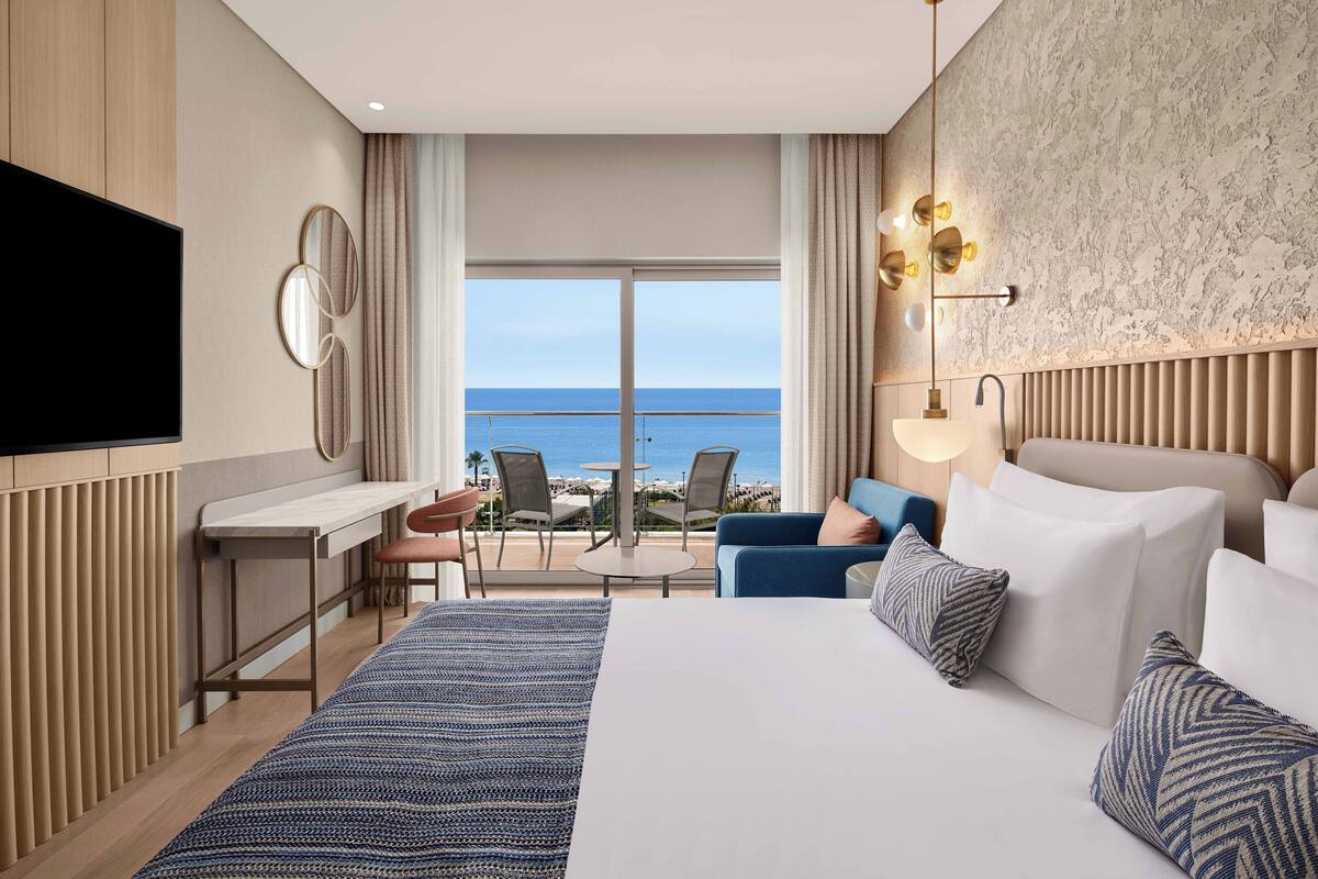 A hotel room with a bed, a desk, a chair, and a view of the sea.