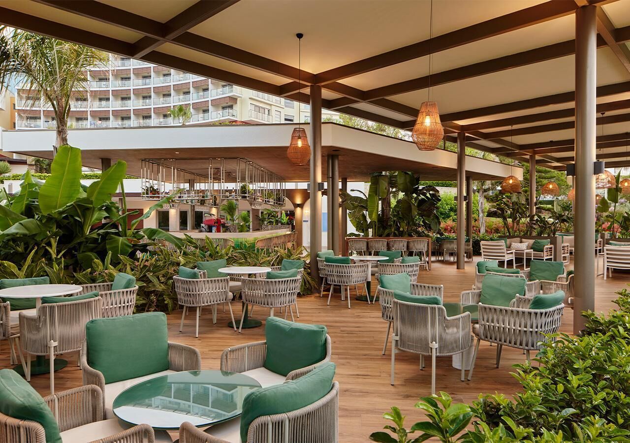An outdoor dining area with wicker furniture and green cushions, surrounded by plants and a view of a building.