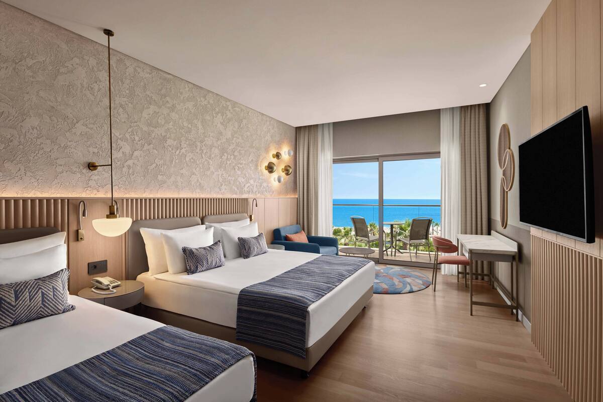 A hotel room with two beds, a TV, a desk, and a balcony with a view of the sea.
