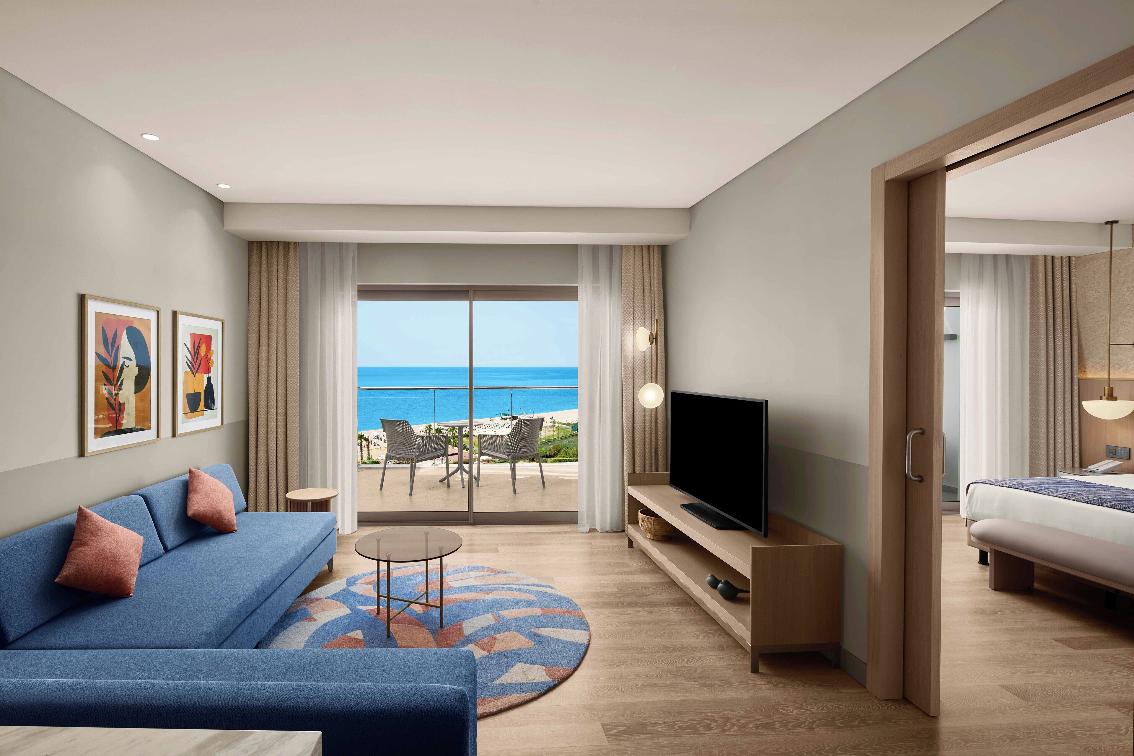 A modern hotel room with a sofa, a TV, a bed, and a balcony with a view of the sea.