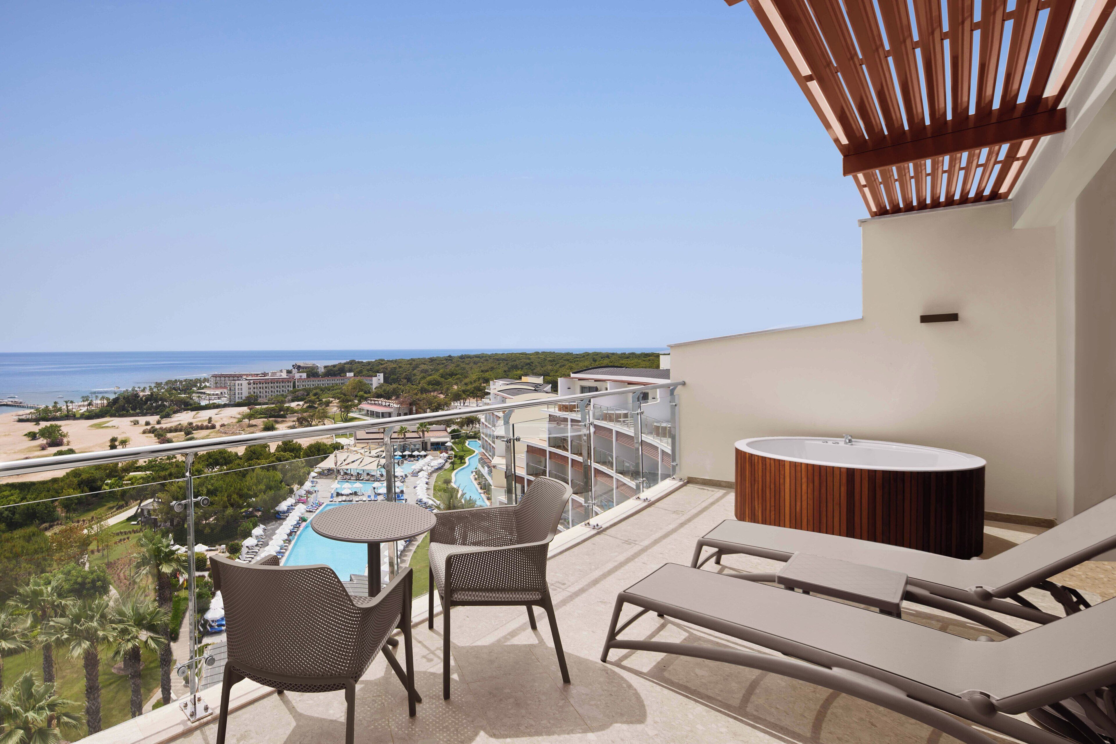 A balcony with a view of a beach and a pool. There are lounge chairs and a table.
