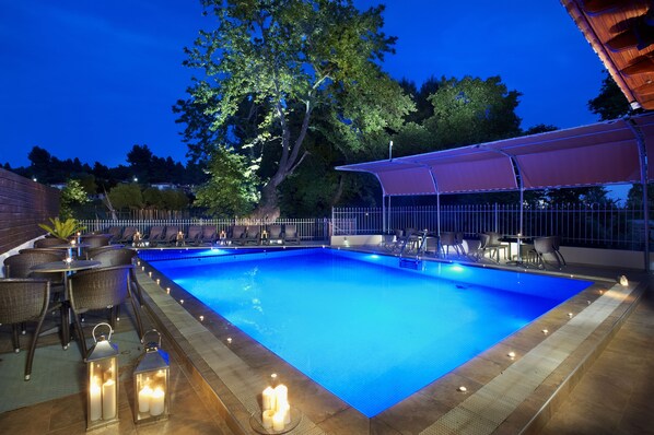 A rectangular swimming pool with a blue interior, surrounded by a covered seating area with chairs and tables, and a brick wall in the background. - 4 you Hotel (Polygyros)