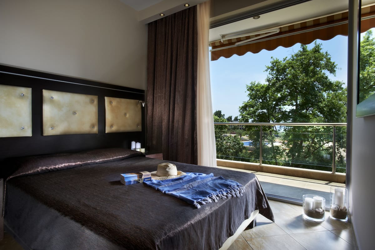 A hotel room with a large bed, a view of trees through the window, and a balcony with a railing.