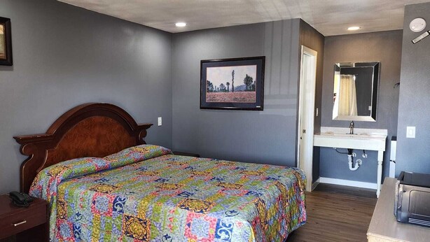 A bedroom with a wooden bed, a bedside table, a nightstand, a dresser, a bathroom with a sink and mirror, and two framed pictures on the wall. - Economy Inn Victorville (Victorville)
