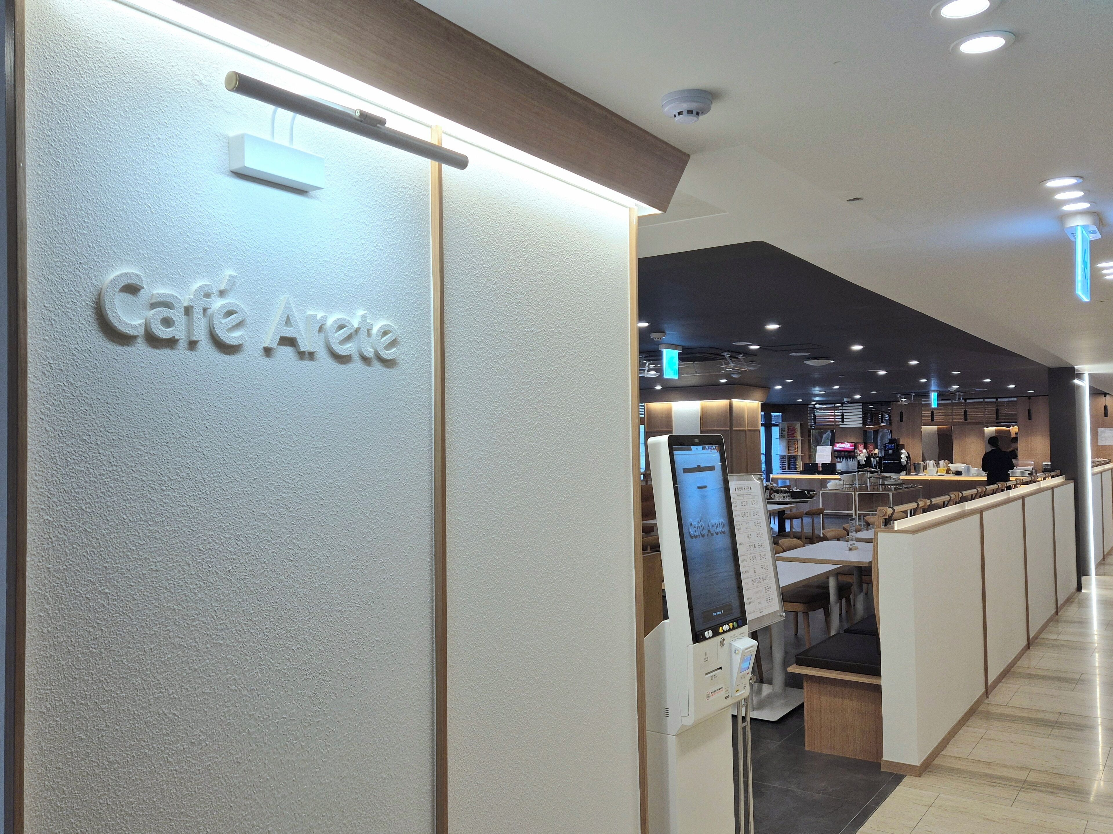 L'intérieur d'un café moderne, avec une enseigne indiquant « Café Arête », un menu numérique et un espace salon avec des tables et des chaises.
