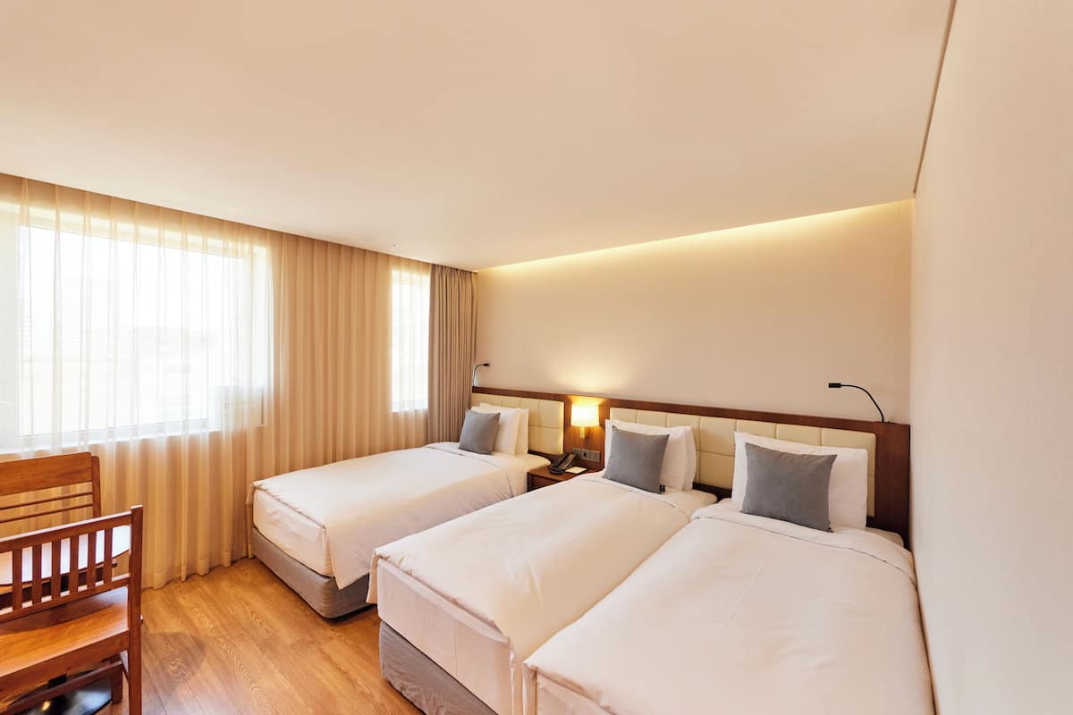 Habitación de hotel con dos camas individuales, una silla de madera, un escritorio y una ventana con cortinas.