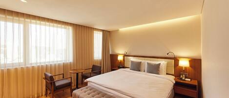 A hotel room with a large bed, a bedside table with a lamp, a chair, a small table, and a window with curtains.