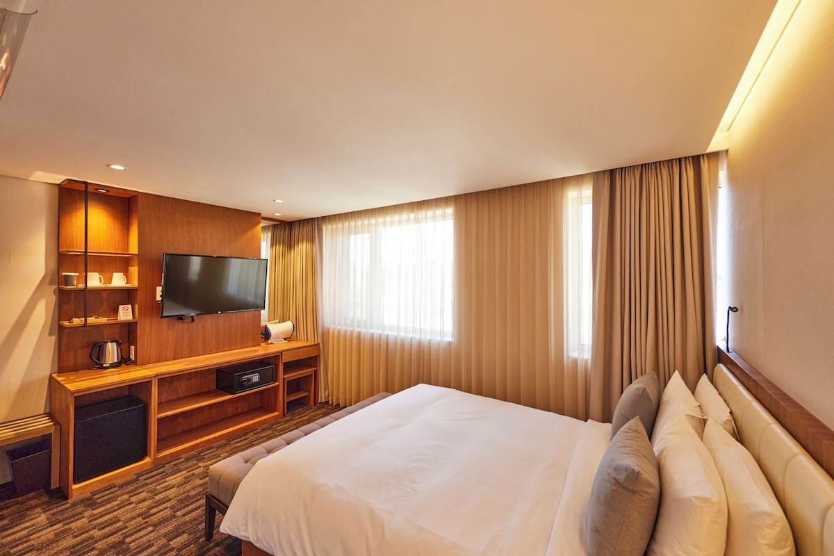 A hotel room with a large bed, a flat-screen TV, a wooden desk, and a minibar.