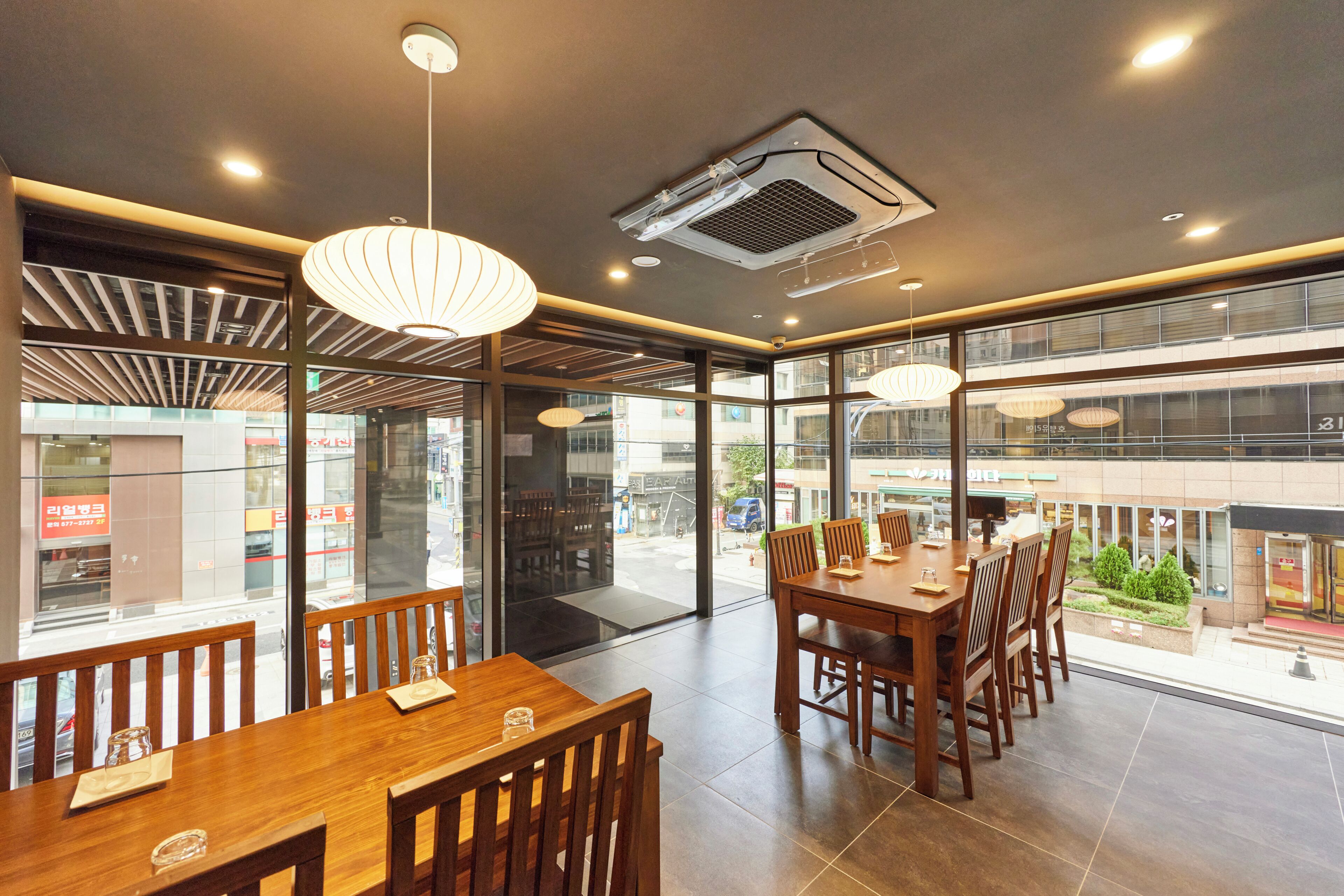 A modern restaurant interior with wooden tables and chairs, pendant lighting, and large windows offering a view of the street.