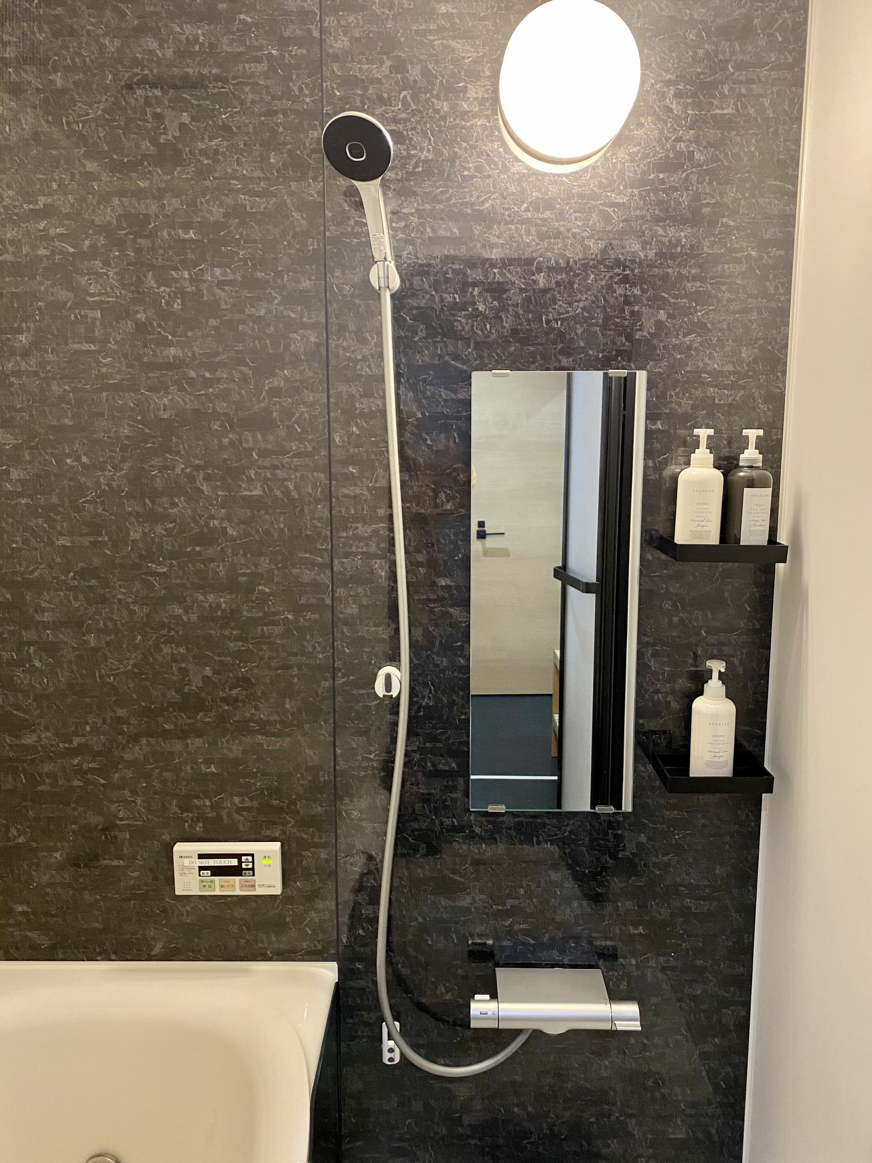 A modern bathroom with a black marble wall, a handheld showerhead, a built-in toilet, a mirror, and two bottles of lotion on a shelf.