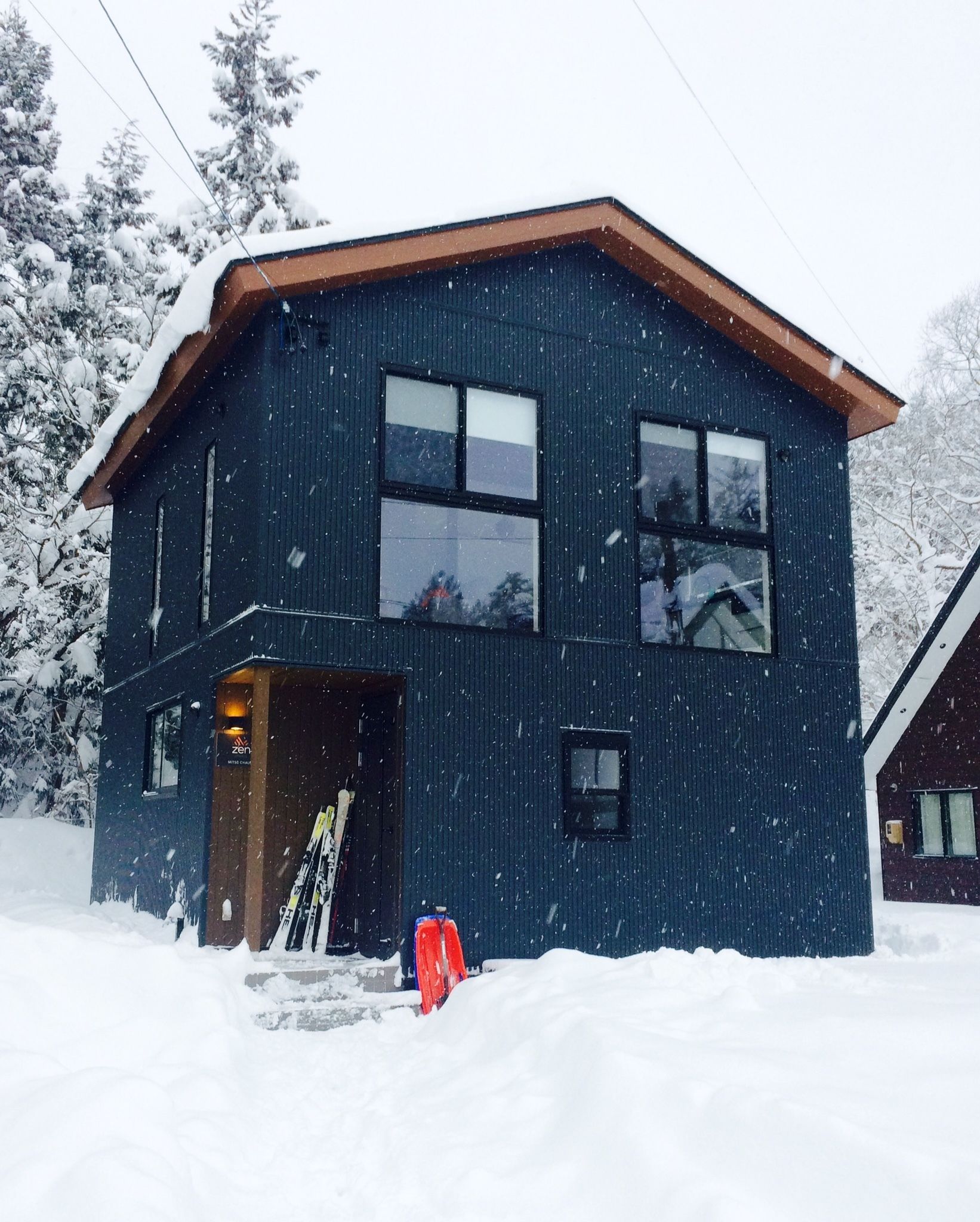 A modern two-story house with a dark exterior, large windows, and a sloped roof covered in snow.