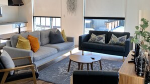 Una sala moderna con un sofá gris, un sofá oscuro y una mesa de centro de madera. Hay una zona de cocina con una estufa y una ventana.