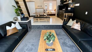 Una sala de estar moderna con sofás de cuero negro, una mesa de centro de madera con una planta y una escalera que lleva a un dormitorio.
