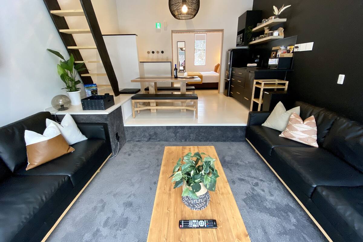 A modern living room with black leather sofas, a wooden coffee table with a plant, and a staircase leading to a bedroom.