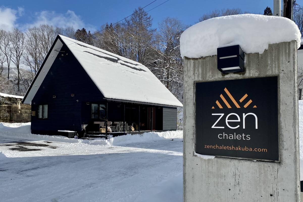 A chalet with a dark exterior and a snow-covered roof, a signboard with the name 'zen chalets' and a website address, and a snowy landscape with leafless trees in the background.