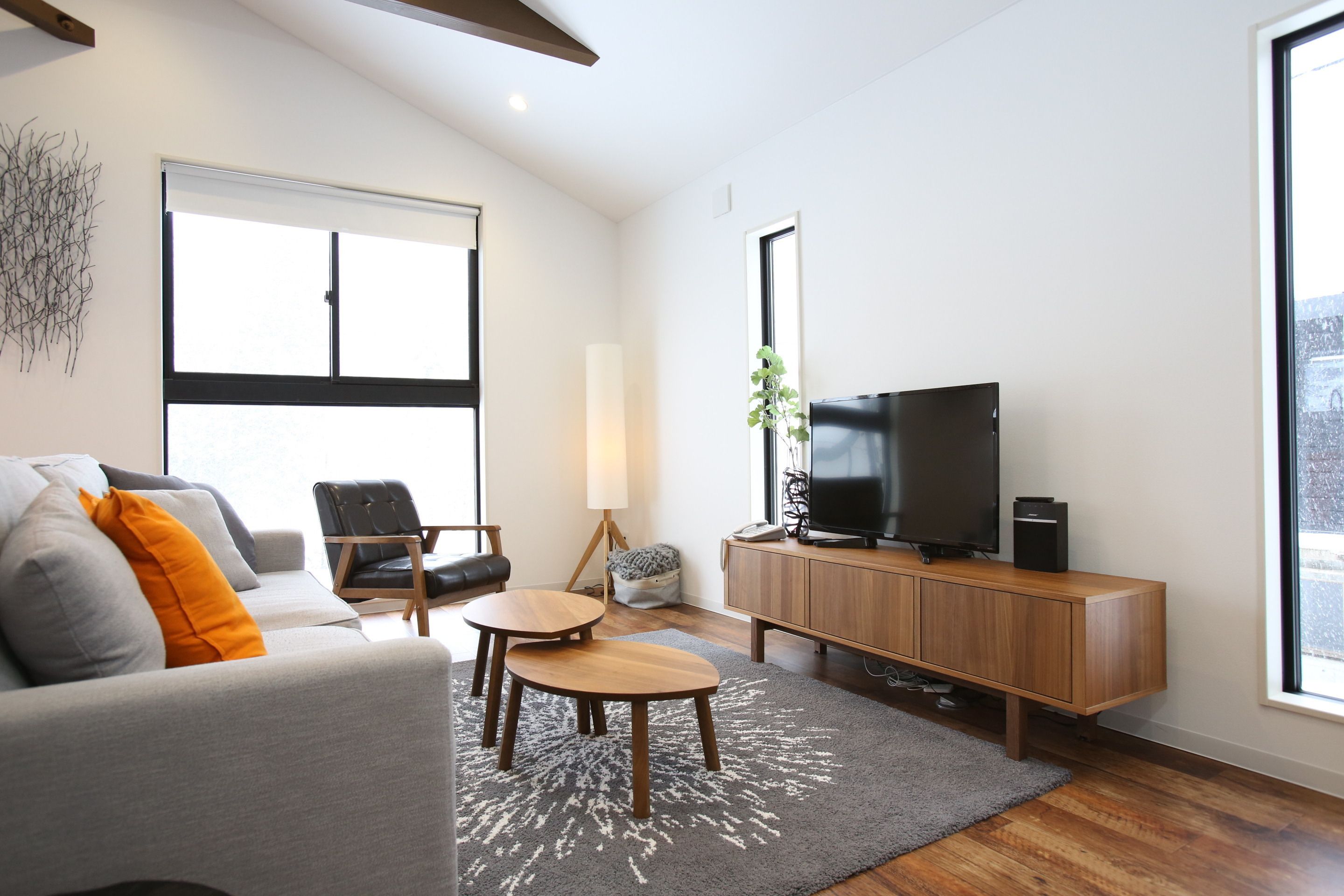 A modern living room with a grey sofa, two wooden side tables, a black leather chair, a wooden TV stand with a flat-screen TV, and a large window offering a view of the outdoors.