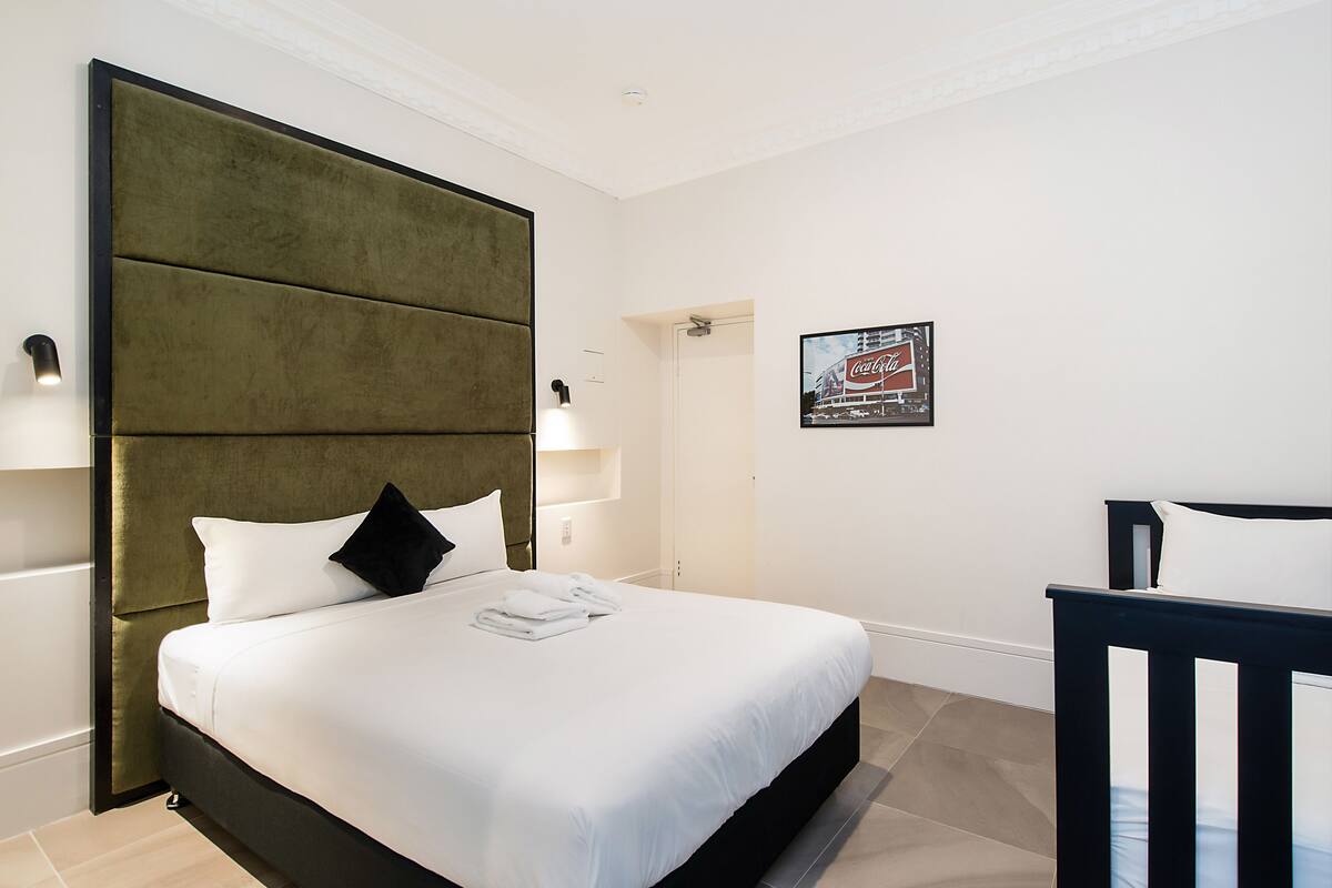 Una cama bien hecha con ropa blanca y un cabecero tapizado en verde, un cuadro en la pared y una silla con estructura negra.