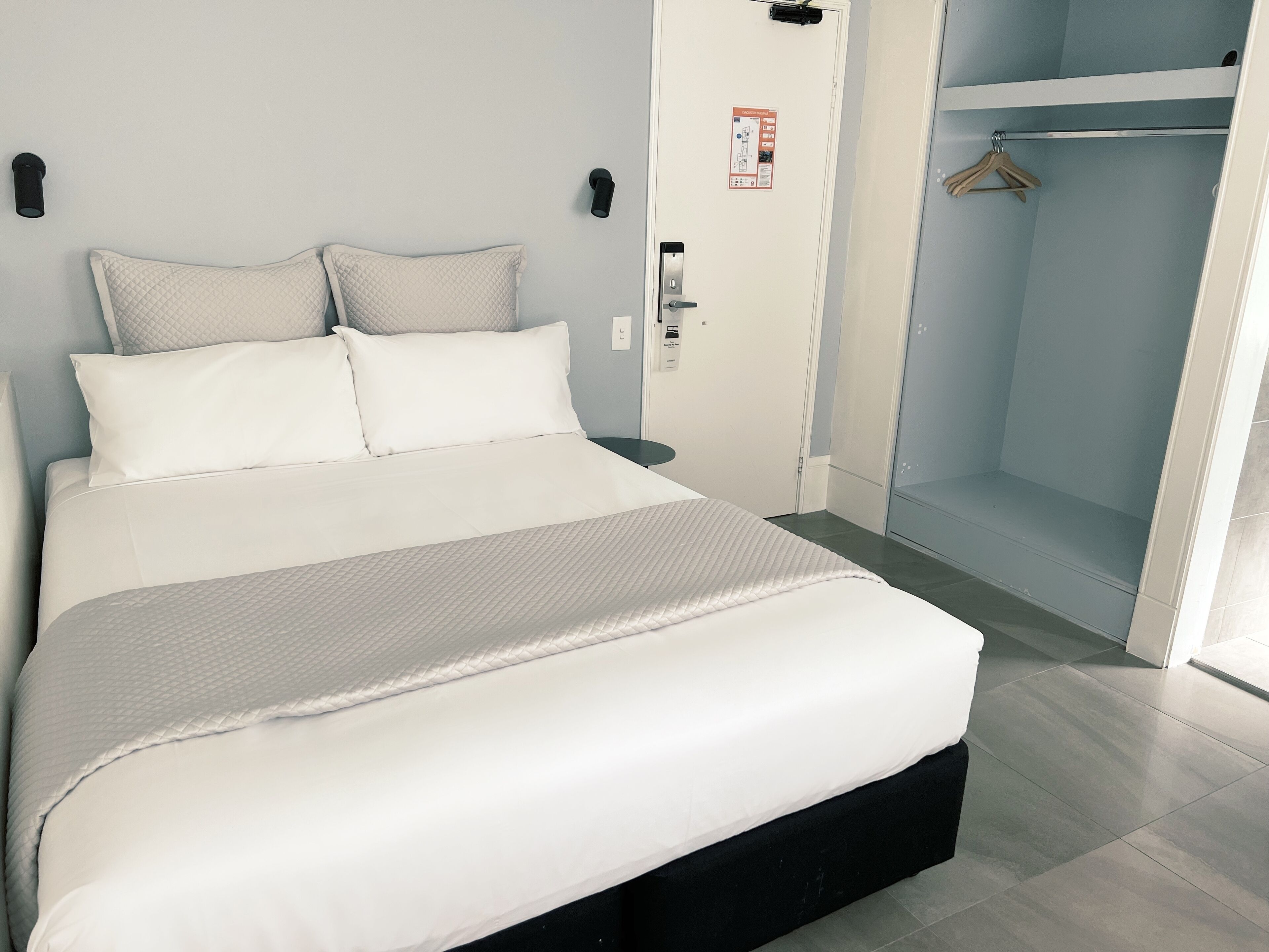 Una cama bien hecha con cabecera negra, pared blanca con señal de seguridad contra incendios y vestidor con barra para colgar ropa.