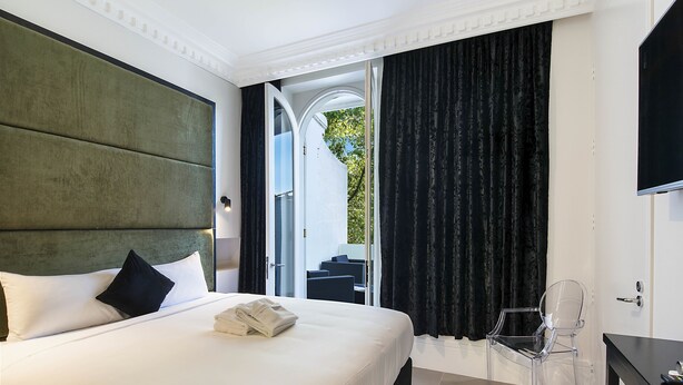 A hotel room with a large bed, a clear chair, a desk, and a view through an open door. - Sydney Boutique Hotel (Darlinghurst)
