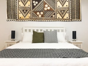 Un dormitorio con cama, cabecera blanca, dos mesitas de noche con lámparas y un tapiz decorativo en la pared.