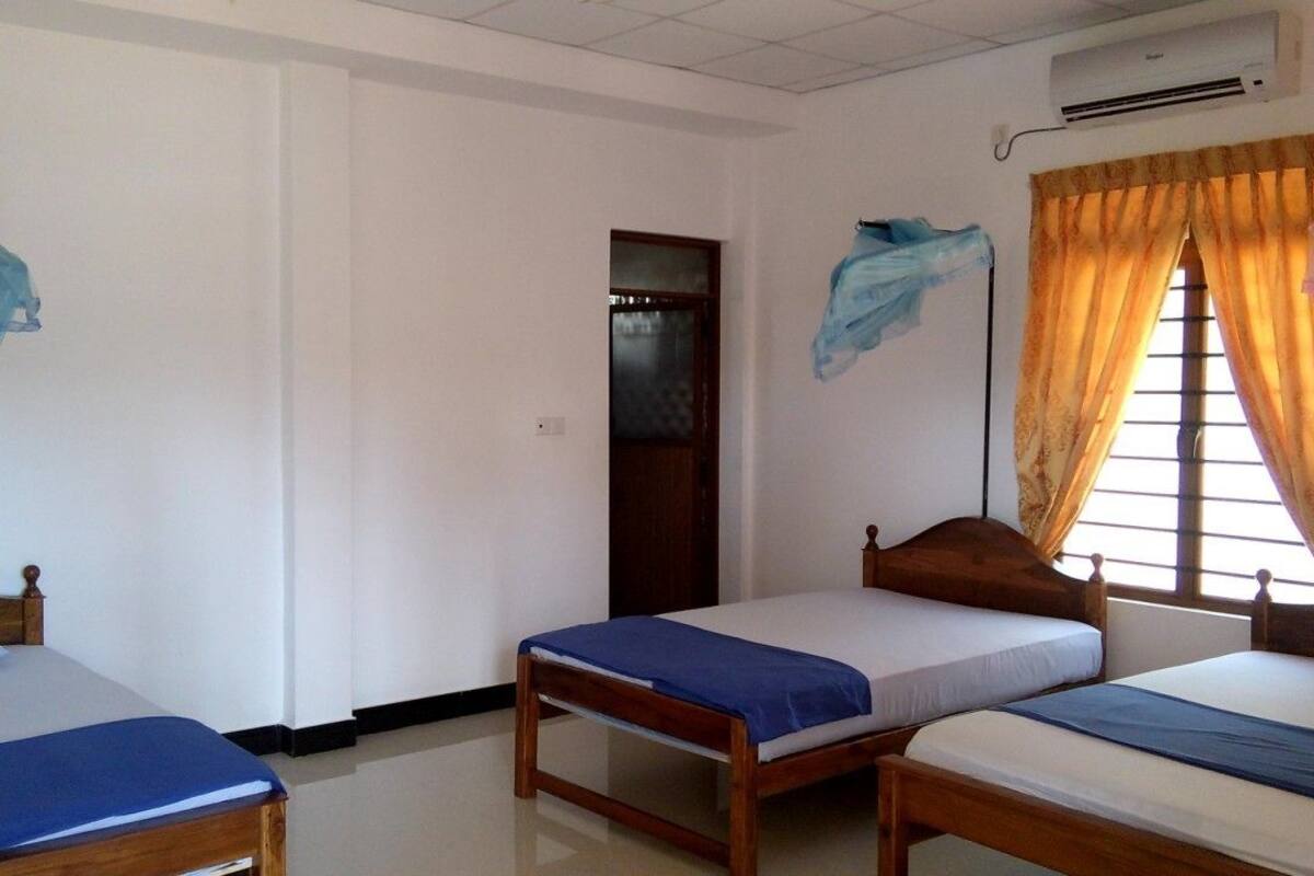 A room with three wooden beds, each with a blue bedspread. There is an air conditioning unit on the wall, and a window with curtains.