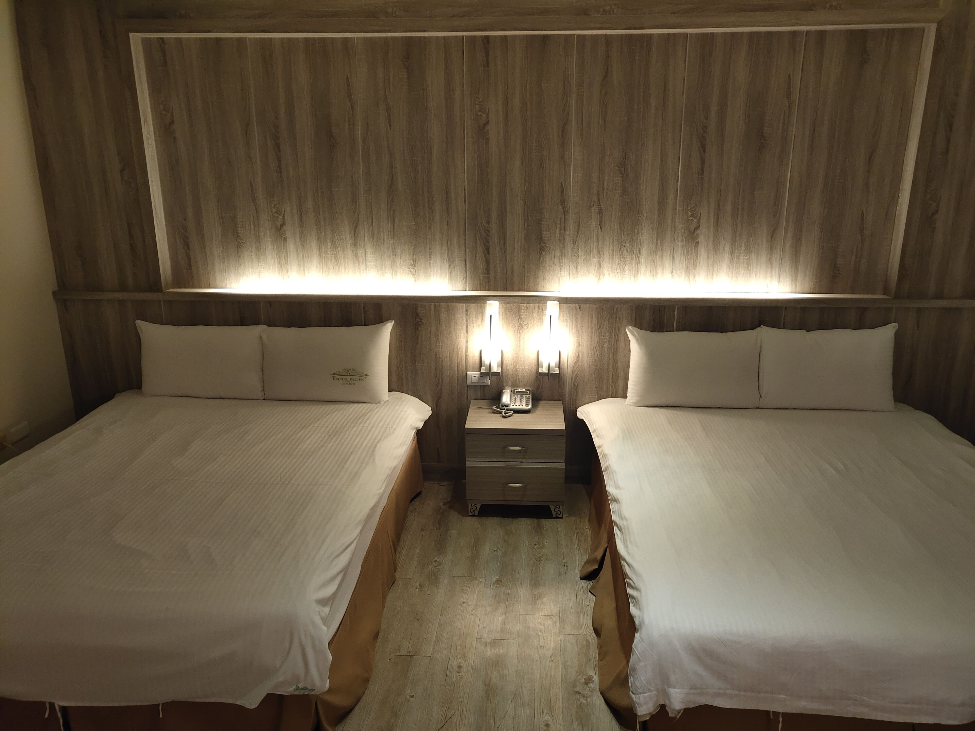 Een hotelkamer met twee bedden, een houten hoofdbord, bedlampen en een klein tafeltje met een telefoon.