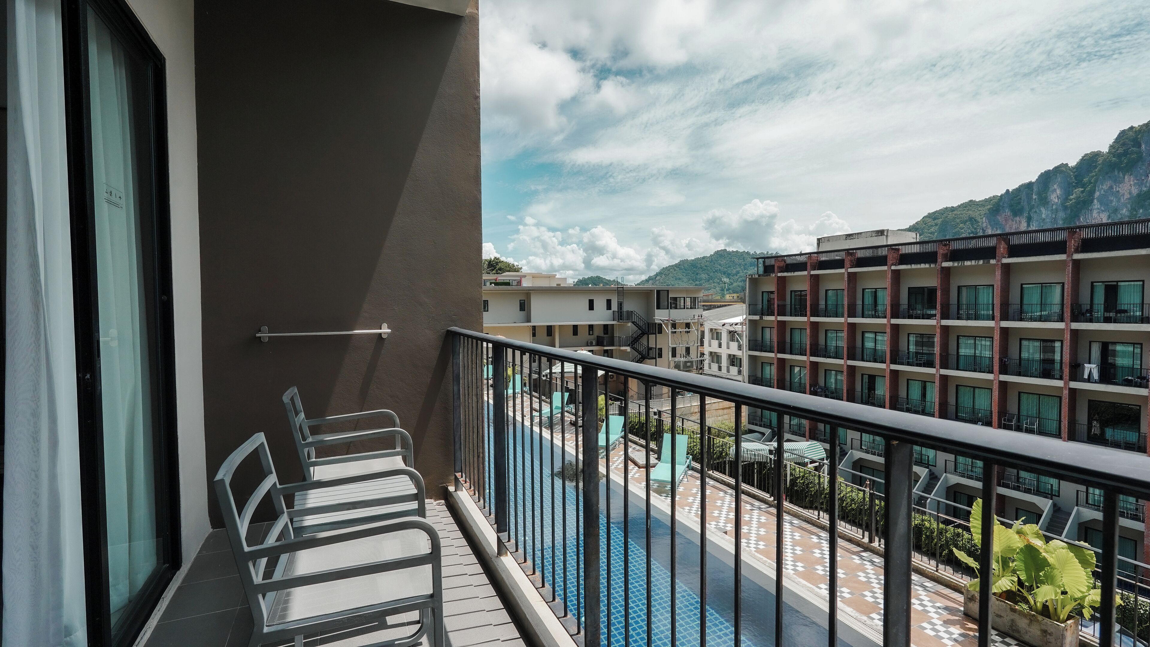 A balcony with a view of a swimming pool, a building with multiple floors, and a mountainous landscape in the background.