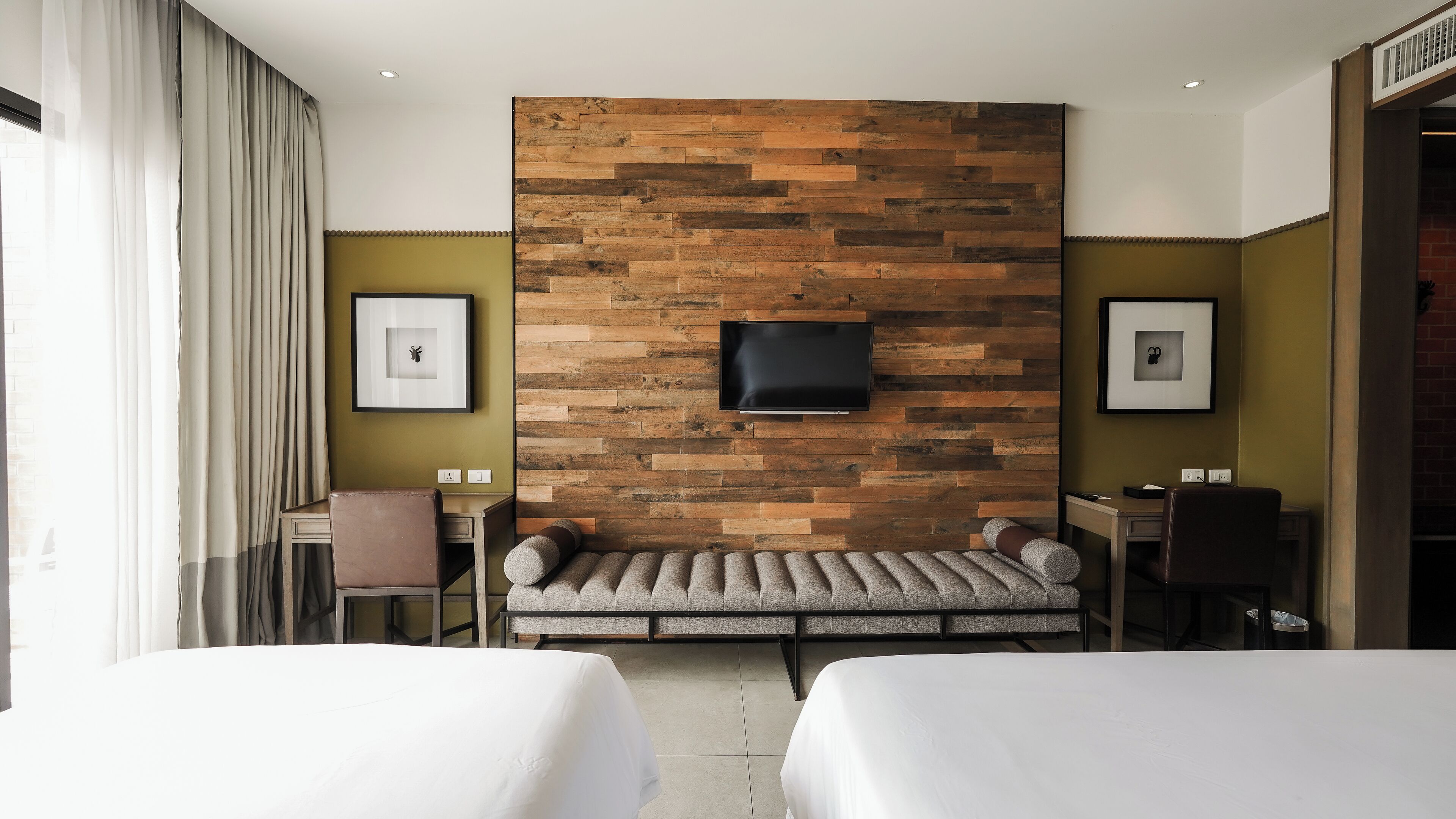 A hotel room with two beds, a sofa bed, a desk, a chair, a TV mounted on a wooden wall, and two framed pictures.