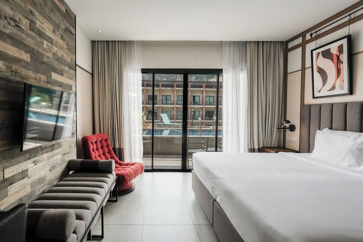 Una habitación de hotel moderna con una cama grande, un sillón rojo, un banco, un televisor de pantalla plana y un balcón con vista a la piscina.