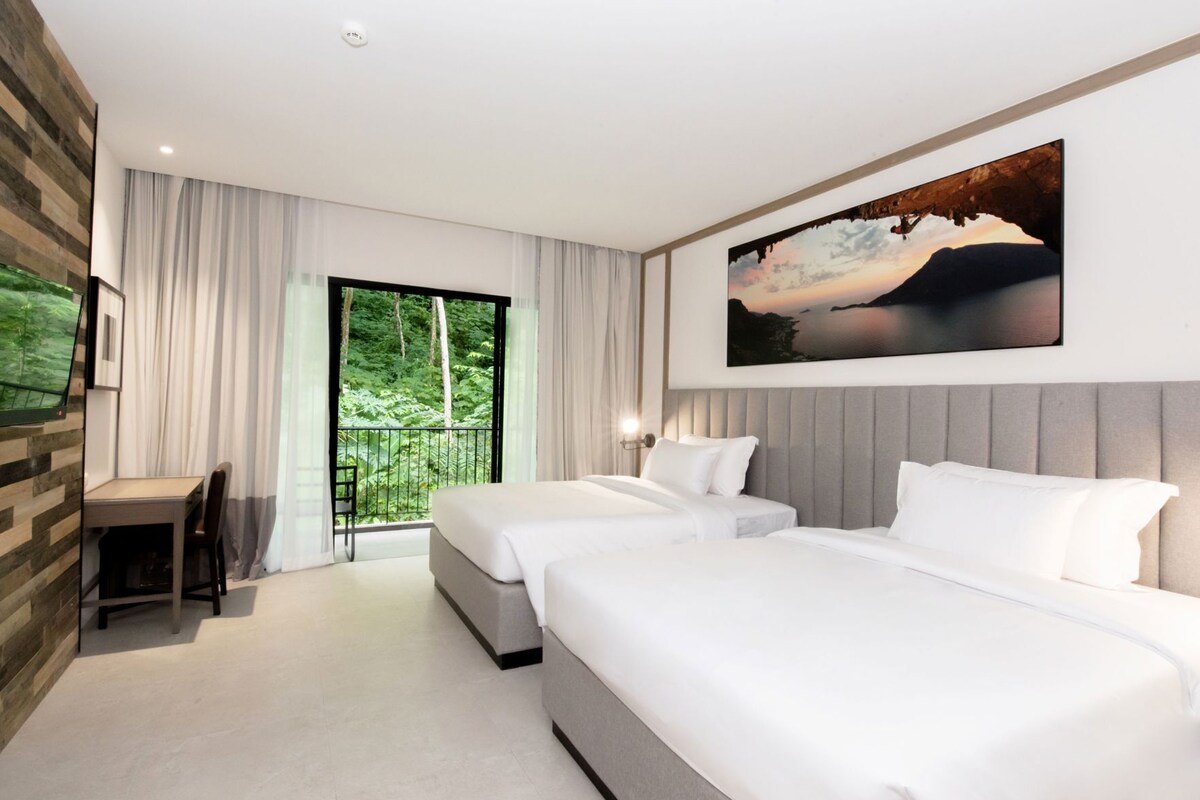 Una habitación de hotel moderna con dos camas, un escritorio, una silla, un balcón con vistas a zonas verdes y un cuadro en la pared.