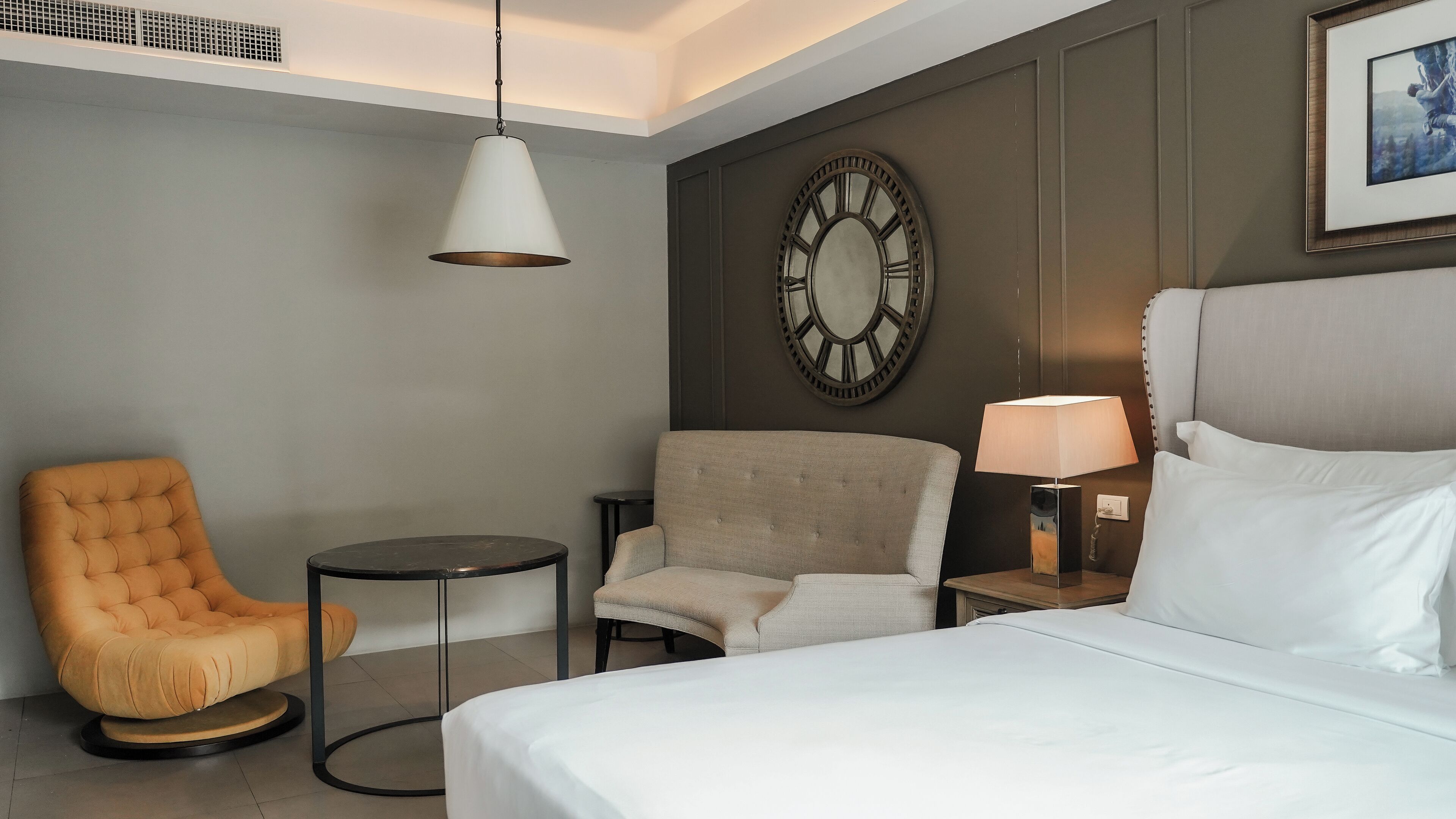 Hotelkamer met een bed, twee fauteuils, een kleine tafel, een wandklok en een hanglamp.