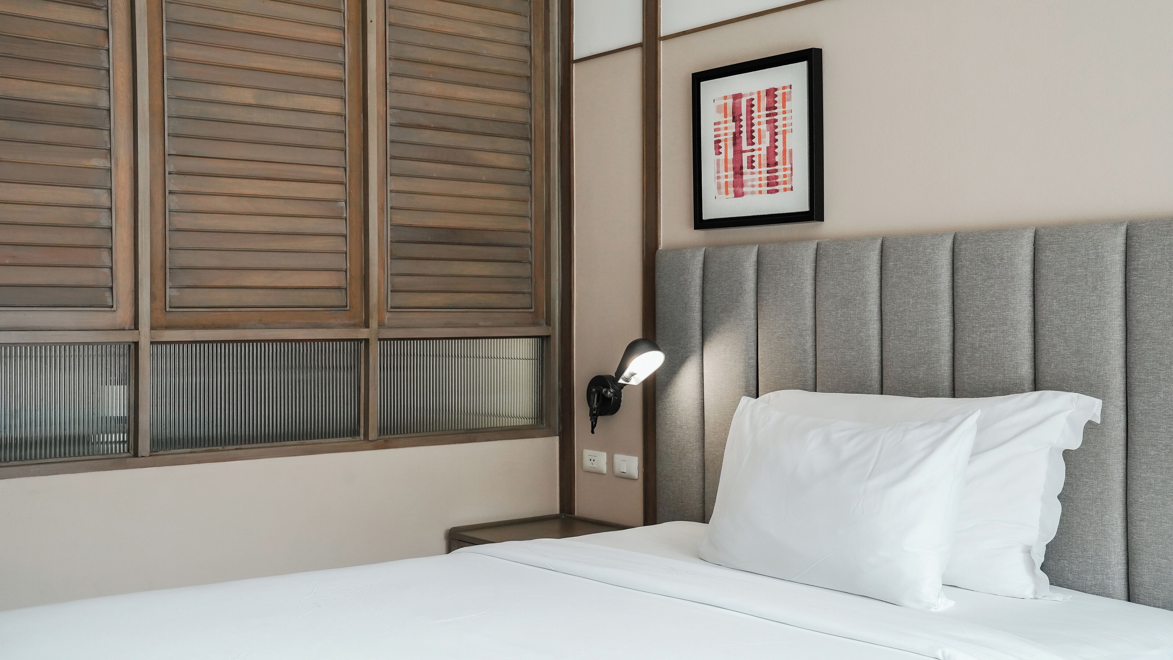 A hotel room with a bed, a nightstand, a wall-mounted lamp, and a window with blinds.