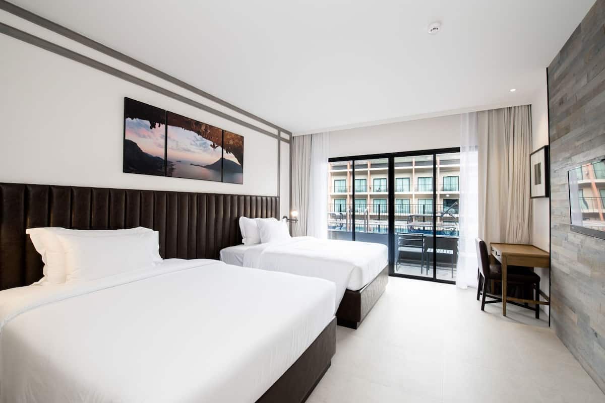 Habitación de hotel con dos camas, un escritorio, una silla, un ventanal con vistas a un edificio y un cuadro en la pared.