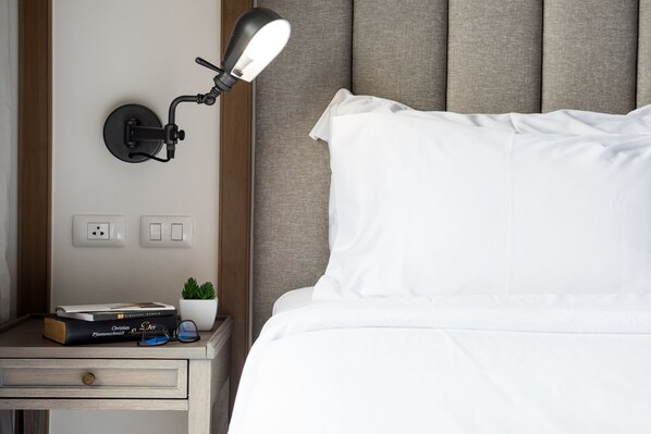 Una cama bien hecha con sábanas blancas, una lámpara de noche, una pequeña planta en maceta y algunos libros en una mesita de noche.