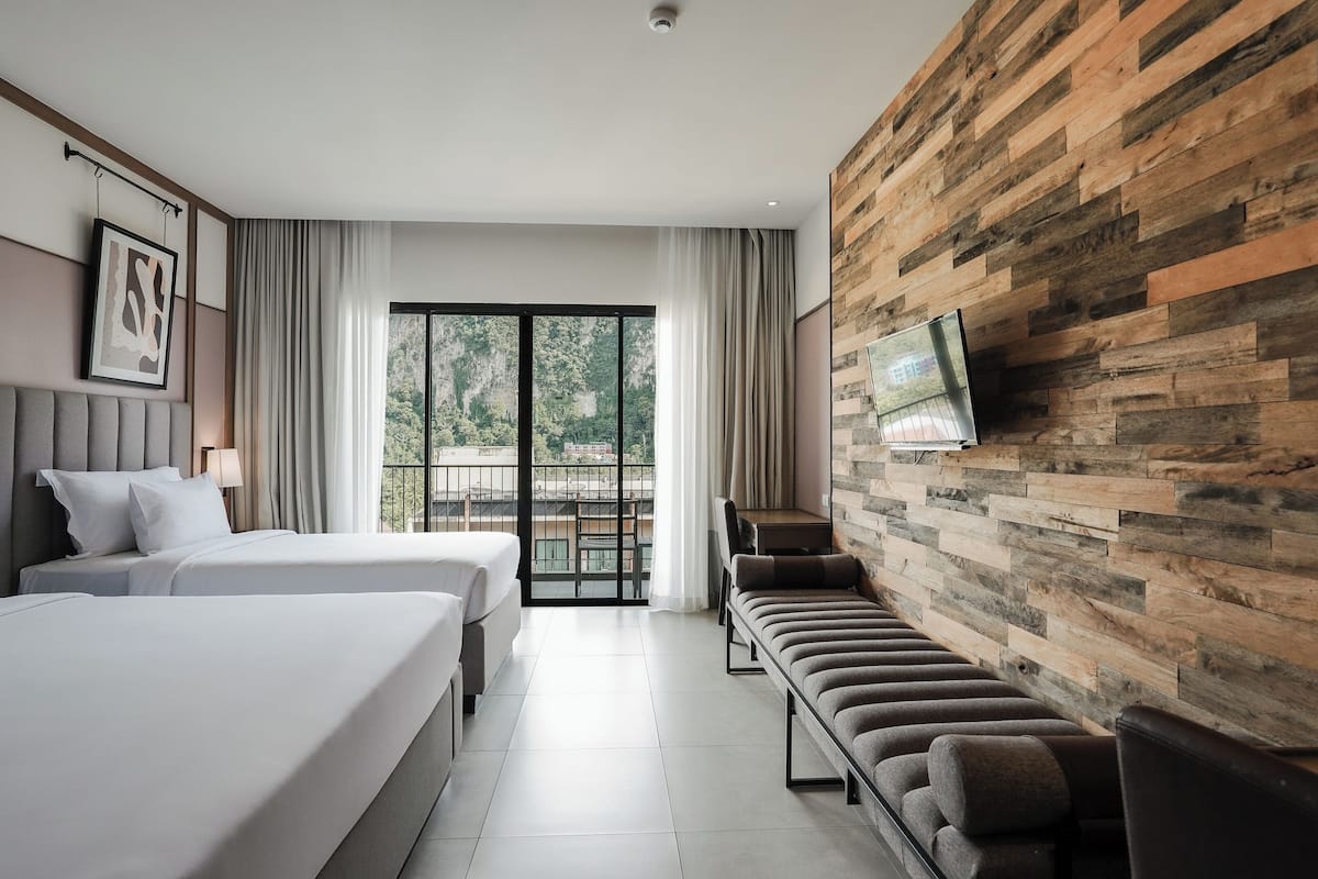 Habitación de hotel con dos camas, un televisor montado en una pared de madera, un balcón con vistas a la vegetación y un área de descanso con sofá y sillas.