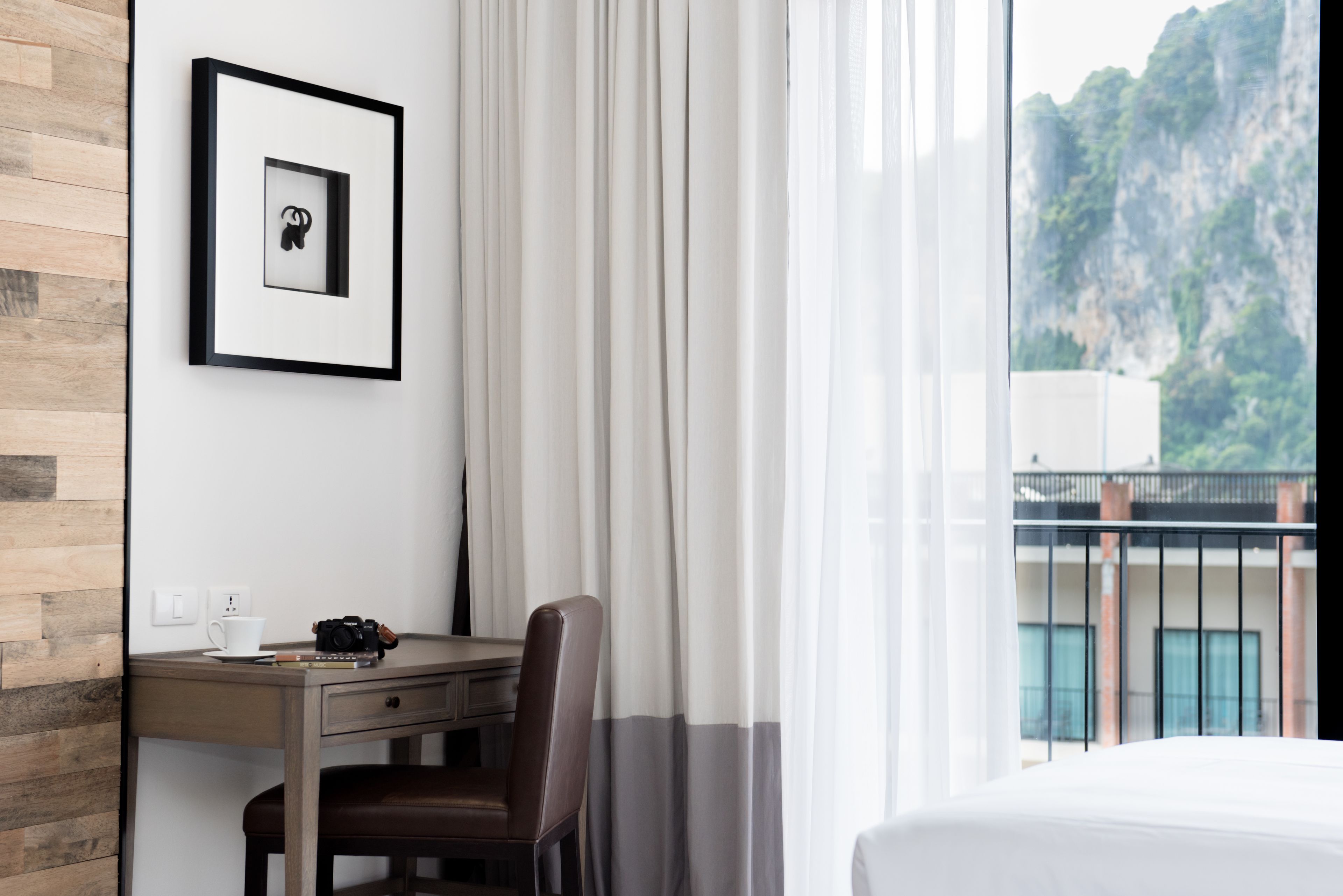 A hotel room with a wooden desk, a chair, a bed, a framed picture on the wall, and a view of a mountainous landscape through the window.
