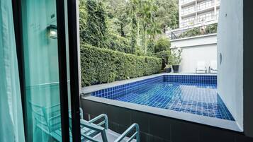Una piscina con azulejos azules, rodeada de follaje y un edificio blanco al fondo.